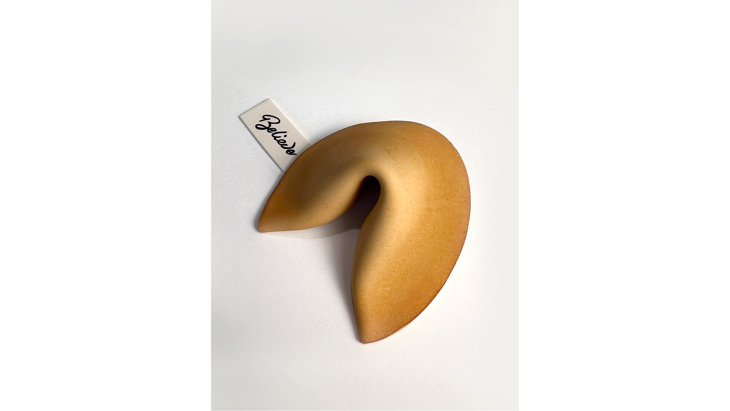 A fortune cookie with a note that says 'Believe' placed next to it on a white surface.