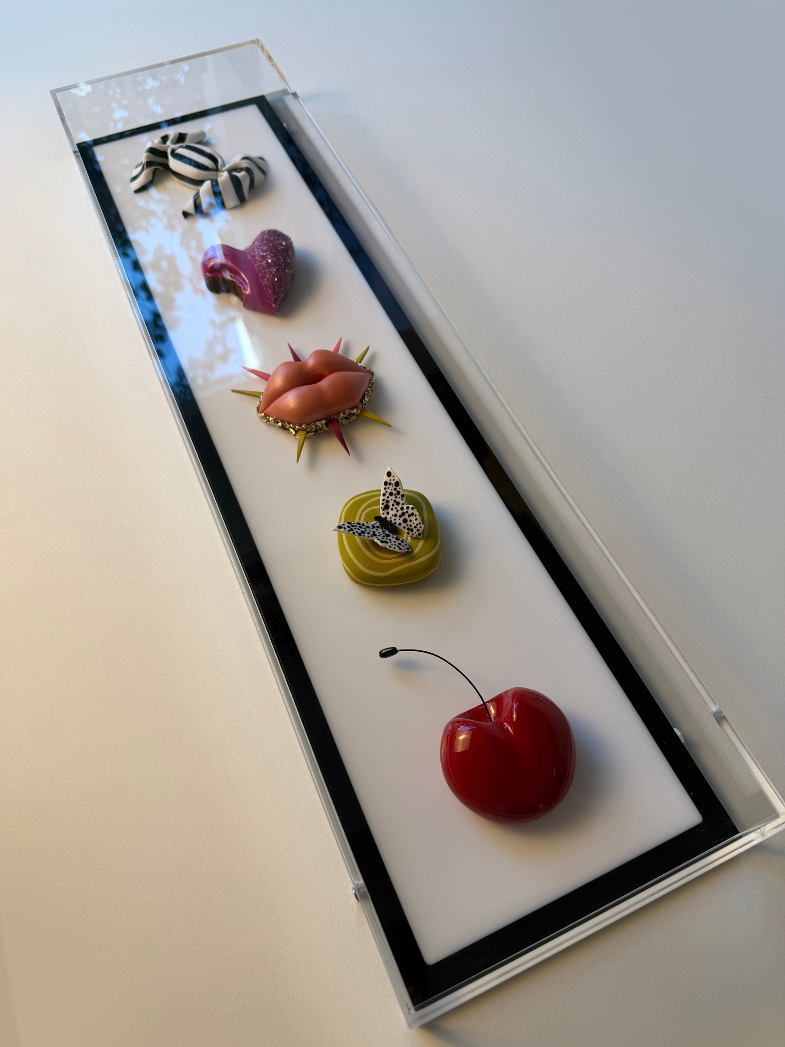 A clear rectangular box with a white interior contains five decorative items: a black and white striped bow, a glittery pink heart, a metallic peach-colored lips, a white butterfly, and a shiny red cherry with a green stem.