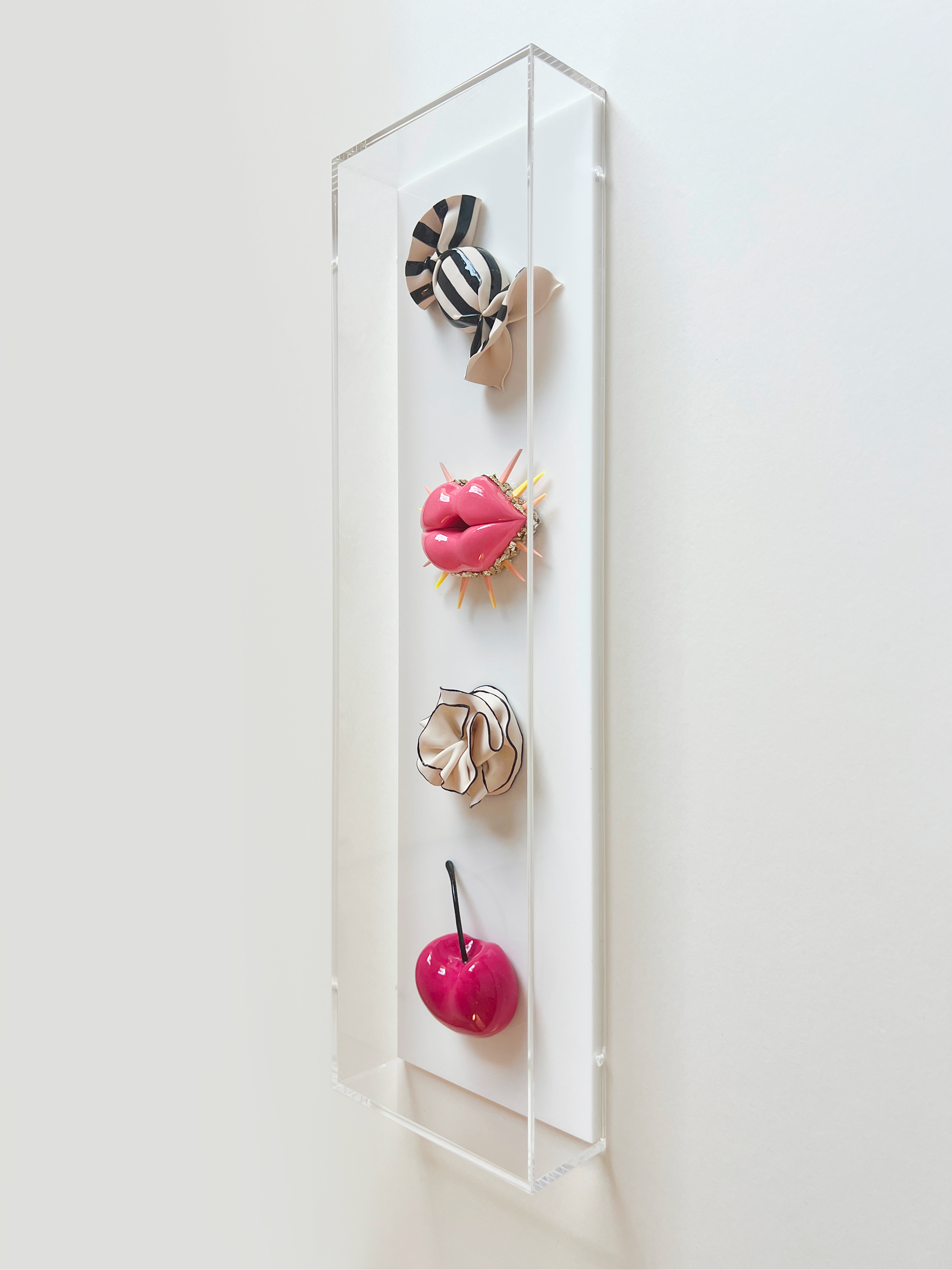 Art installation with a clear acrylic box containing a pink glossy apple with a black straw, a face-shaped object with lips, and a scrunchie with a black and beige pattern, mounted on a white wall.
