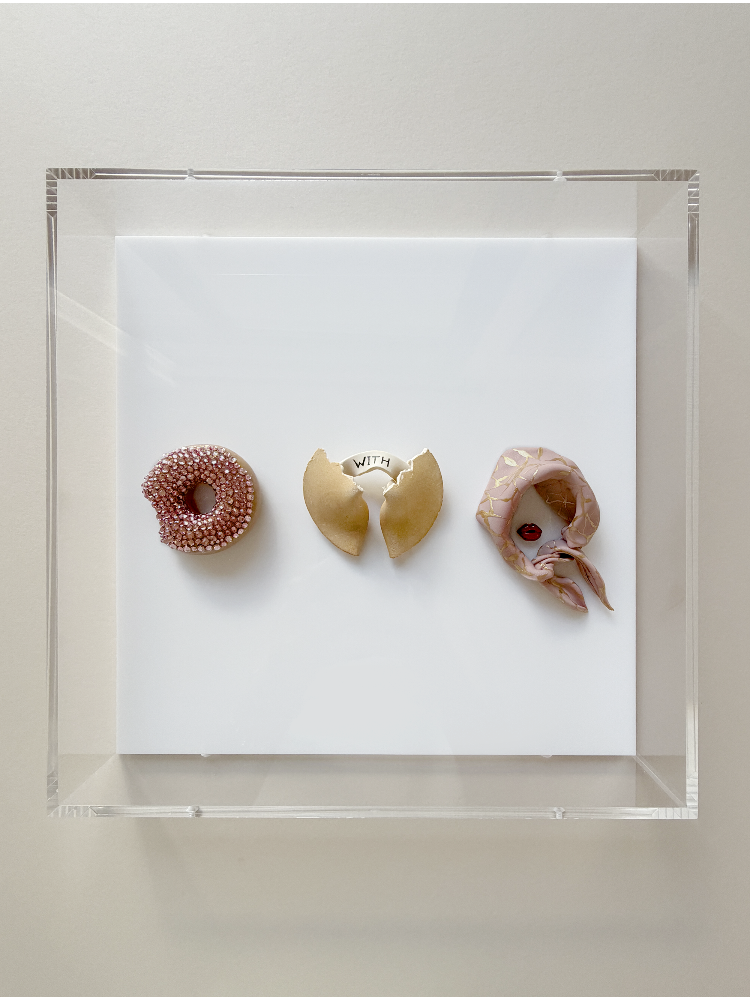 Three decorative cookies on a white surface within a transparent display case. The cookies are shaped like a donut, a butterfly, and a letter Q, decorated with pink, gold, and pink icing with gold accents, and a small red adornment.