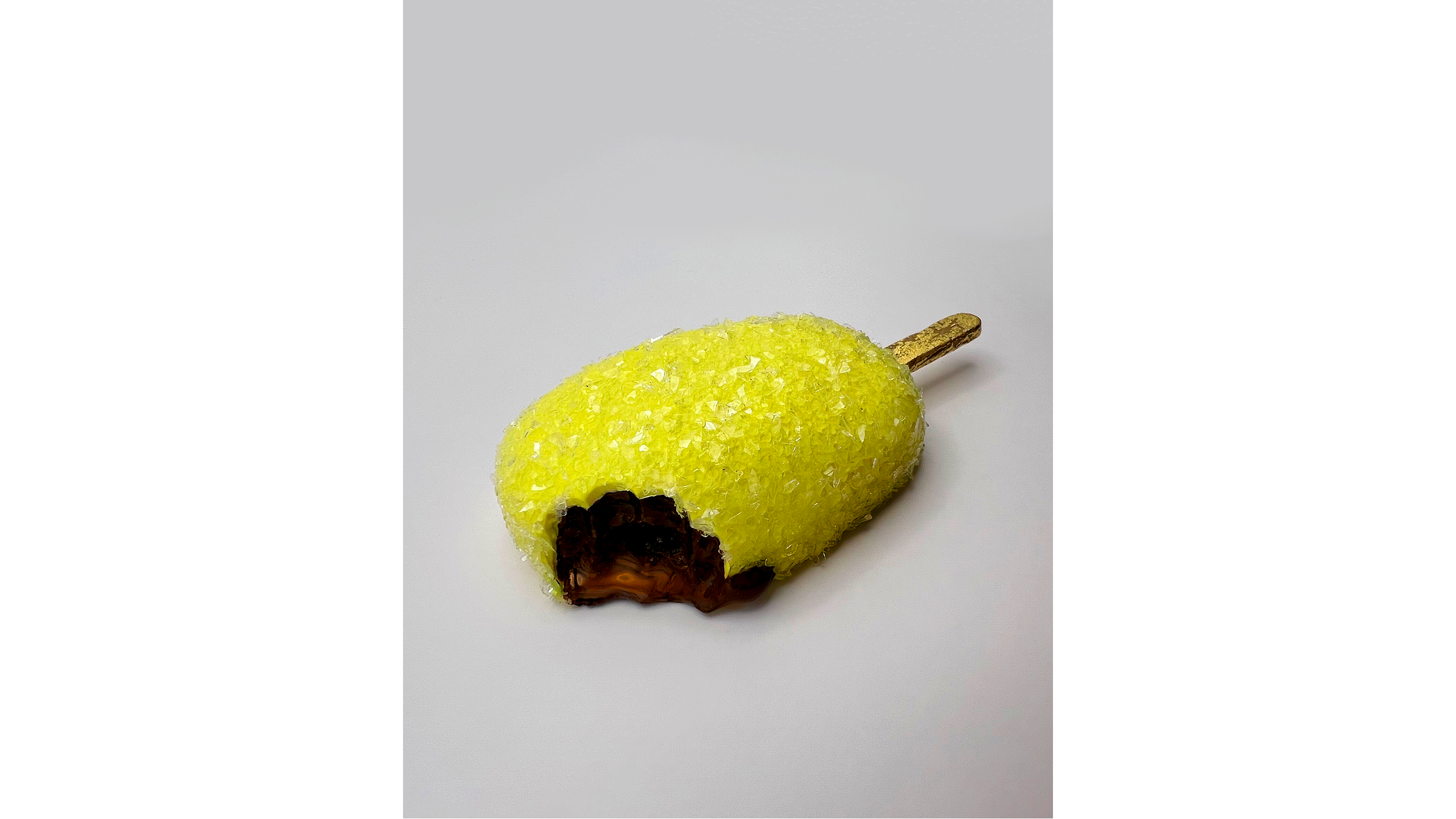 Decorative pear-shaped object made of yellow material with sugar or crystals, filled with dark chocolate at the bottom, and a brown stem on top, placed on a white surface.