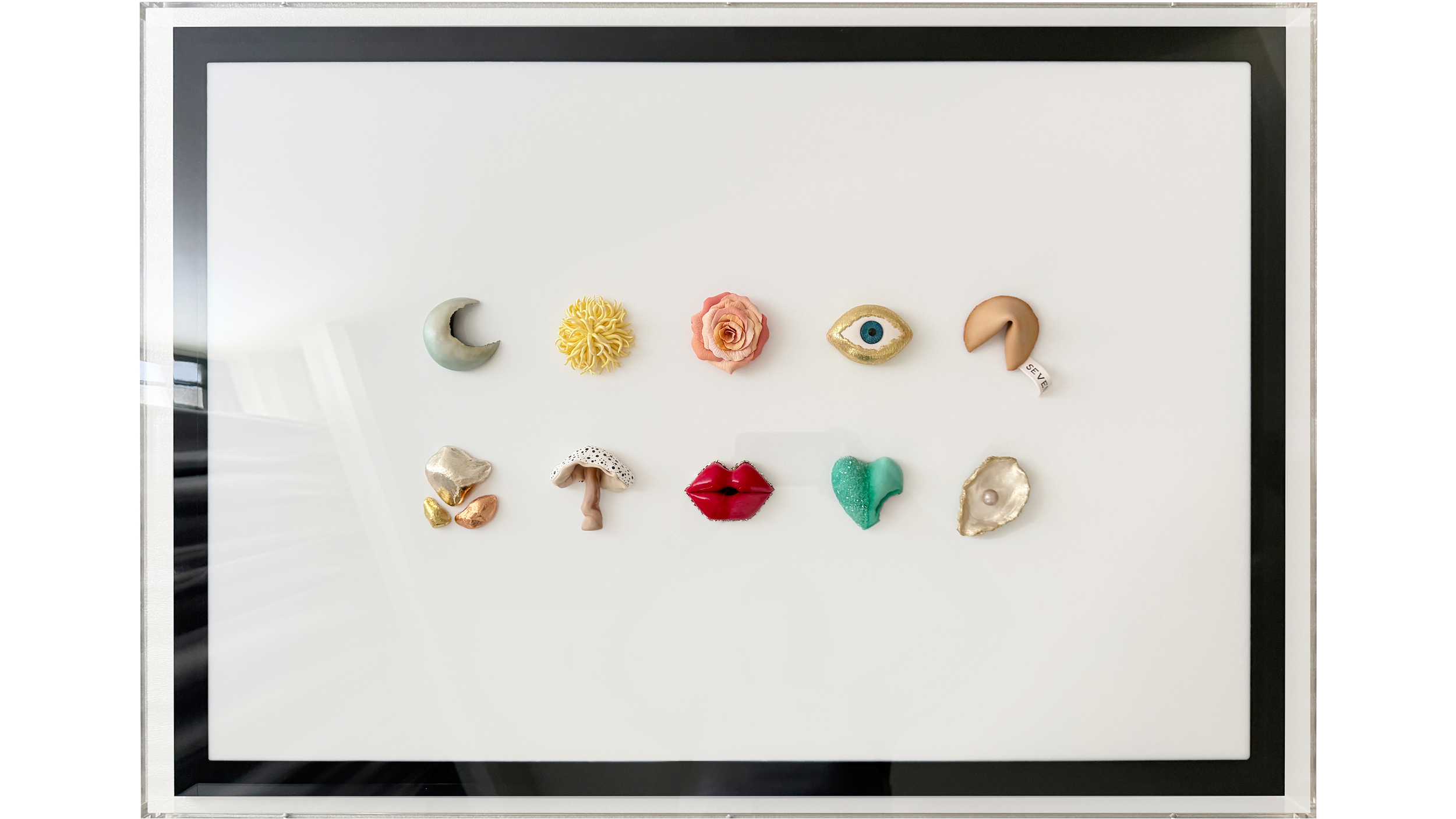 A framed display of various decorative objects, including a crescent moon, orange flower, eye, lips, and other shapes made of different materials.