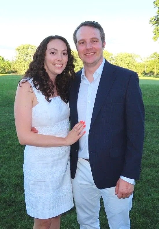 A young couple standing outdoors in a park, smiling at the camera. The woman is wearing a white sleeveless dress, and the man is dressed in a navy blazer, white shirt, and white pants. The background features green grass, trees, and a clear sky.