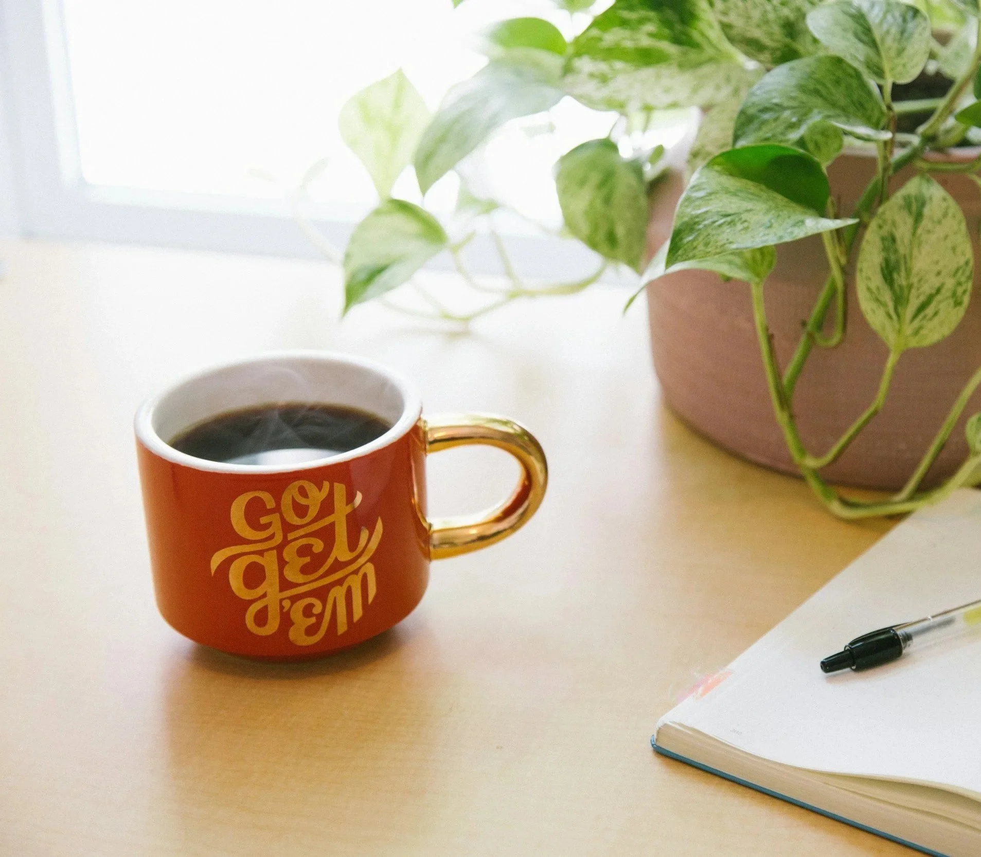 Orange mug with gold handle filled with a dark beverage on a light-colored desk next to a potted plant with green and variegated leaves, a white notebook, and a black pen.