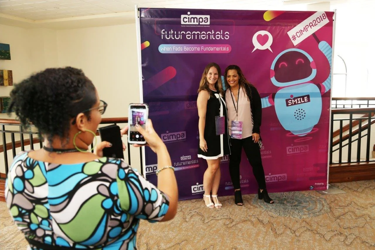 Two women smiling and posing for a photo in front of a purple backdrop at the CIMPA event, with a woman in the foreground taking their picture using her smartphone. The backdrop features a robot and the hashtag #CIMPA2018.