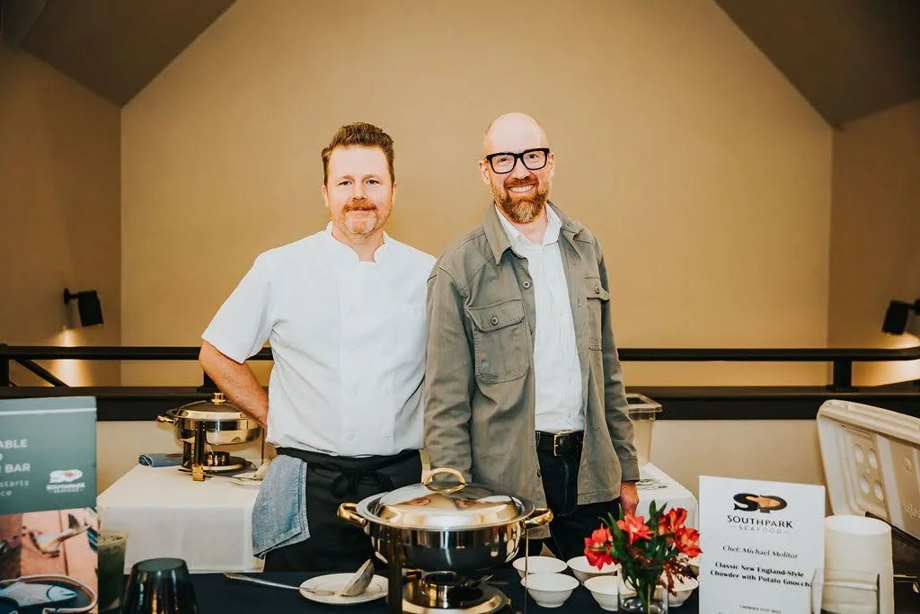 Two men standing behind a buffet table at a seafood event, with chafing dishes, a sign, and a flower arrangement.
