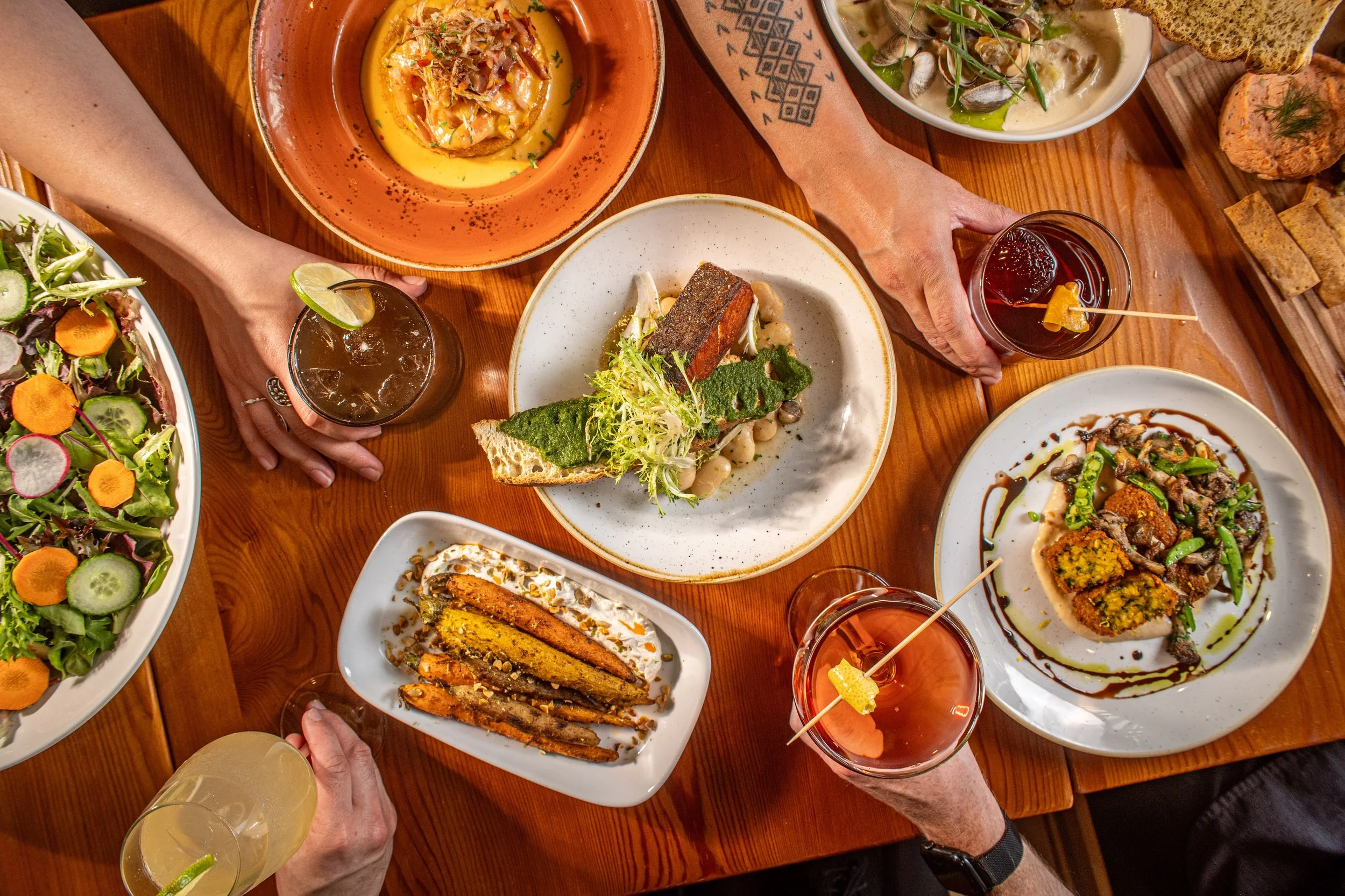 A top-down view of a wooden table with various dishes of food and drinks. Several hands are reaching for glasses with lime wedges, and there are plates of salad, grilled vegetables, and other appetizers.