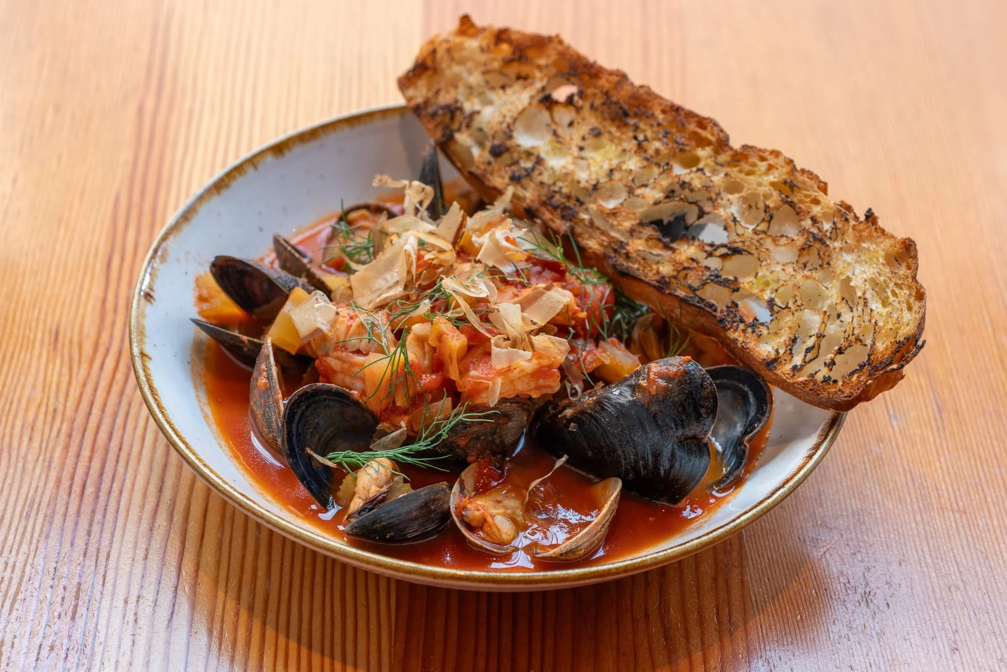 A bowl of seafood stew topped with toasted bread, herbs, and grated cheese, on a wooden table.