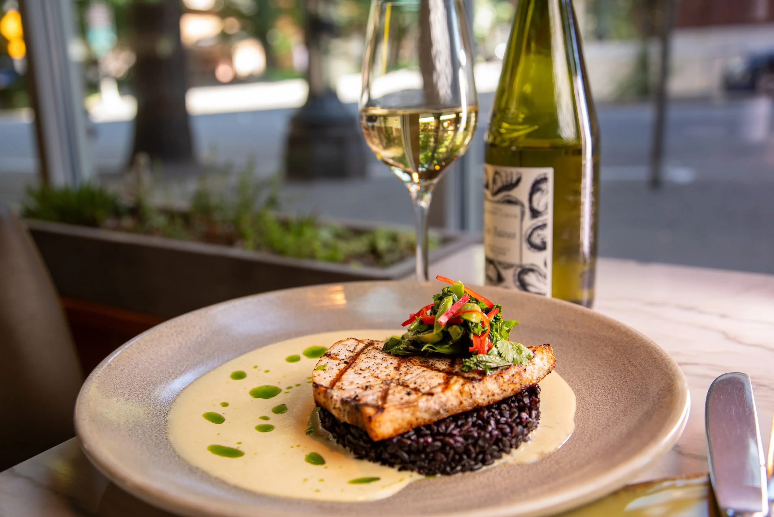 A plated seafood dish with grilled fish topped with a vegetable garnish, served over a bed of black rice with a yellow creamy sauce and green oil drizzle, accompanied by a glass of white wine and a green wine bottle in the background.