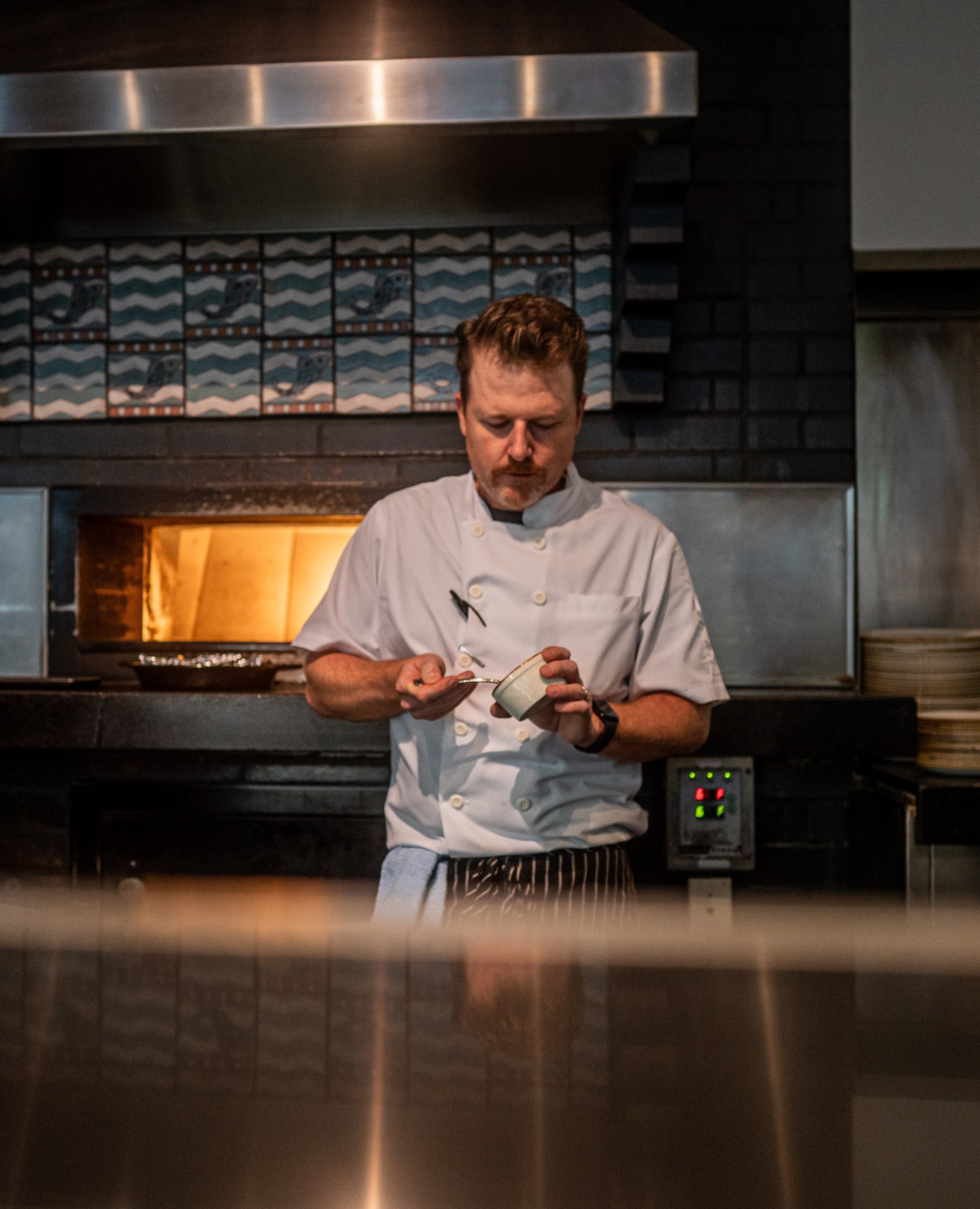 A chef in a white uniform is holding a bowl and a spoon, standing in a commercial kitchen with a black brick wall, decorative tiles, and kitchen appliances.