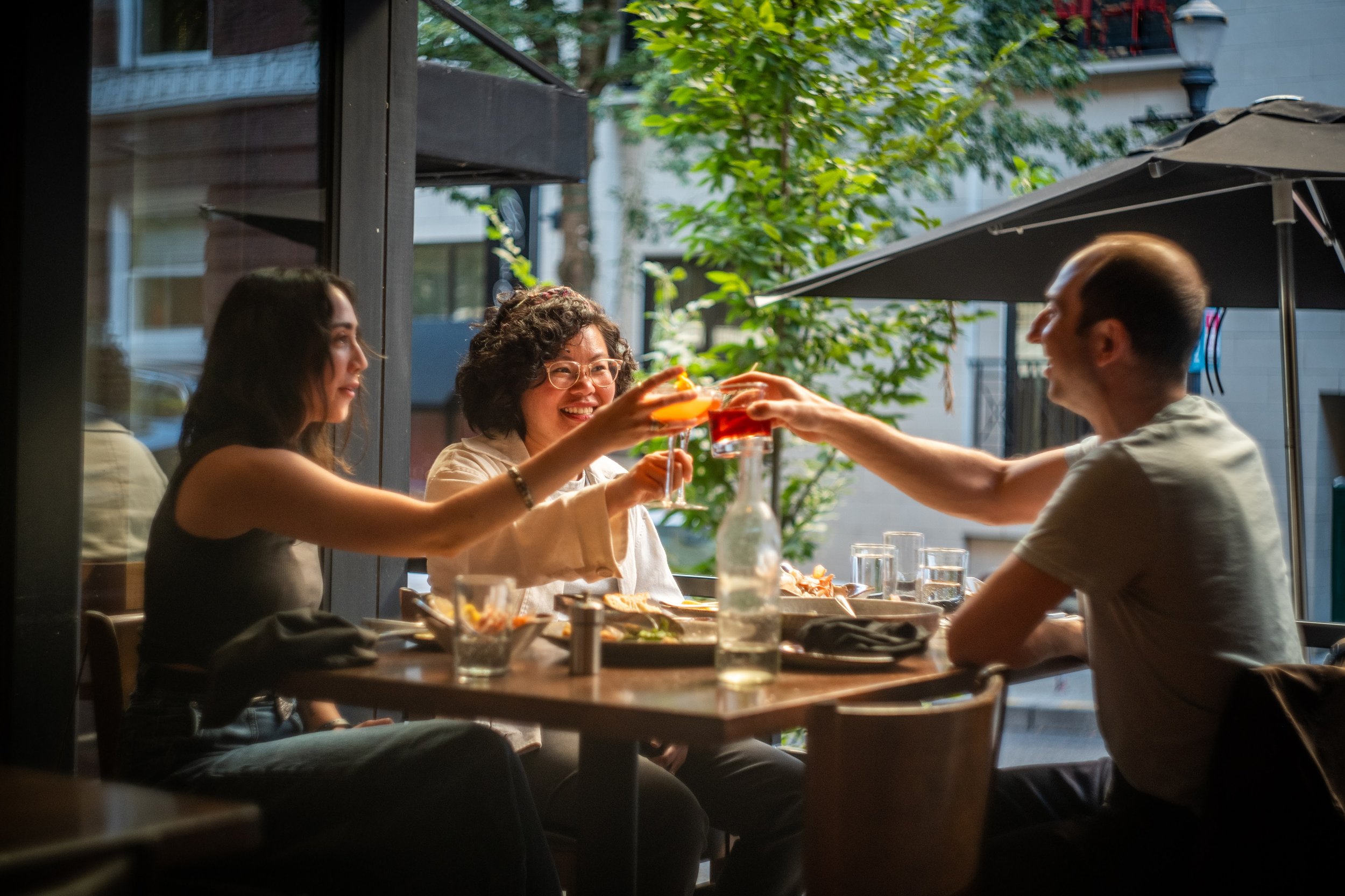 Three people sitting at a table outside a restaurant, sharing drinks and smiling, with greenery and buildings in the background.