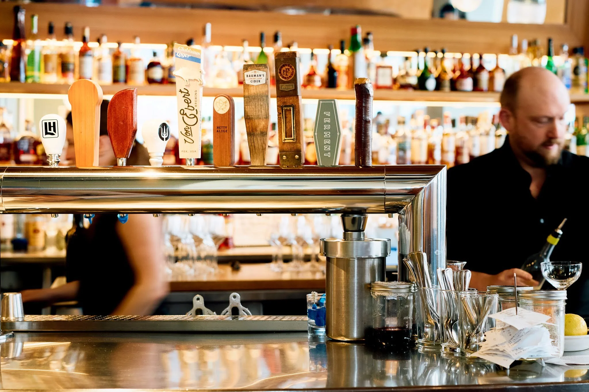 Bar with various beer taps and bottles of alcohol behind the counter, bartender preparing drinks, bar supplies on the counter.