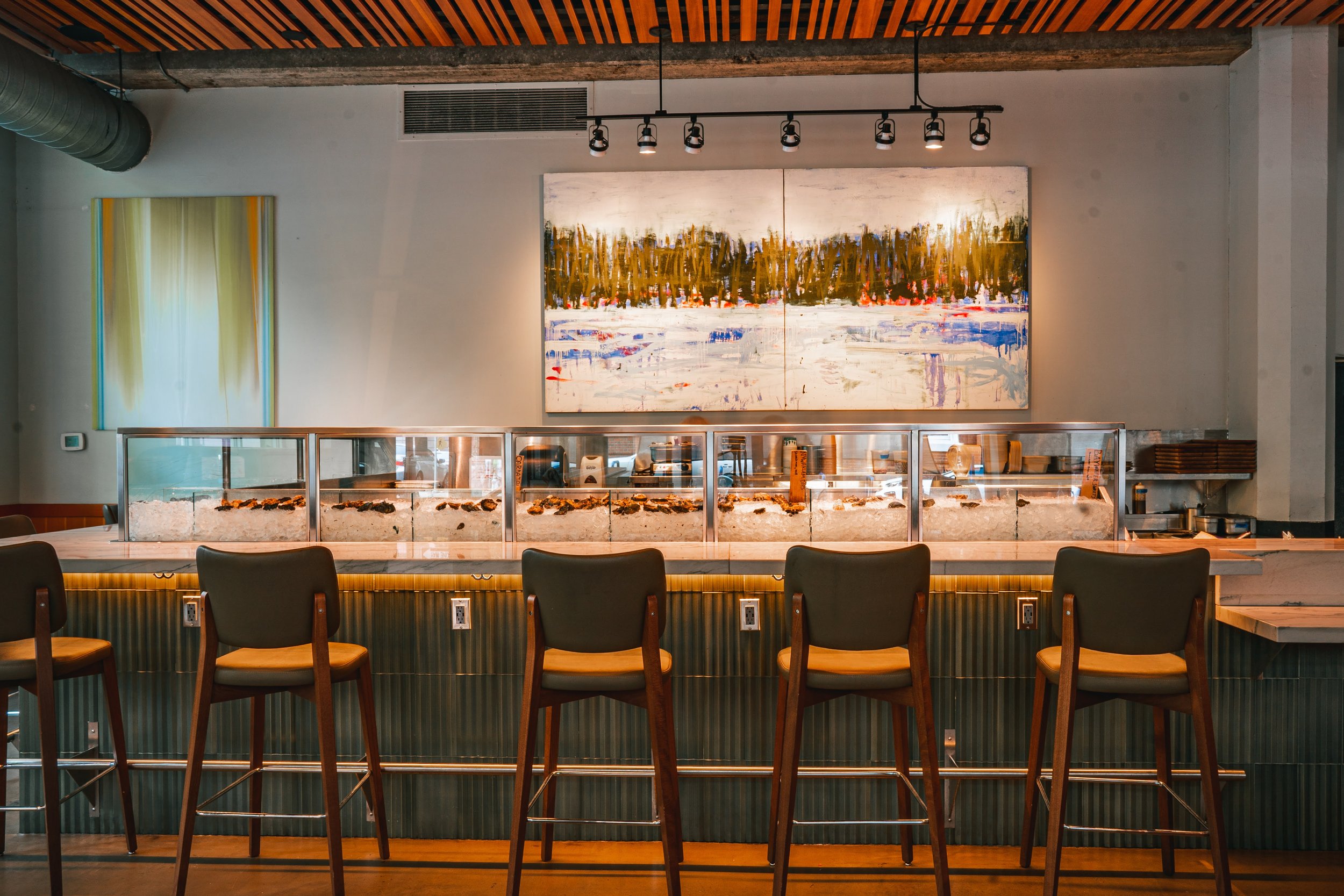 Interior of a modern restaurant with a bar, featuring art on the walls, a wooden ceiling, and chairs in front of the bar counter, with a display of food including sushi and other items.