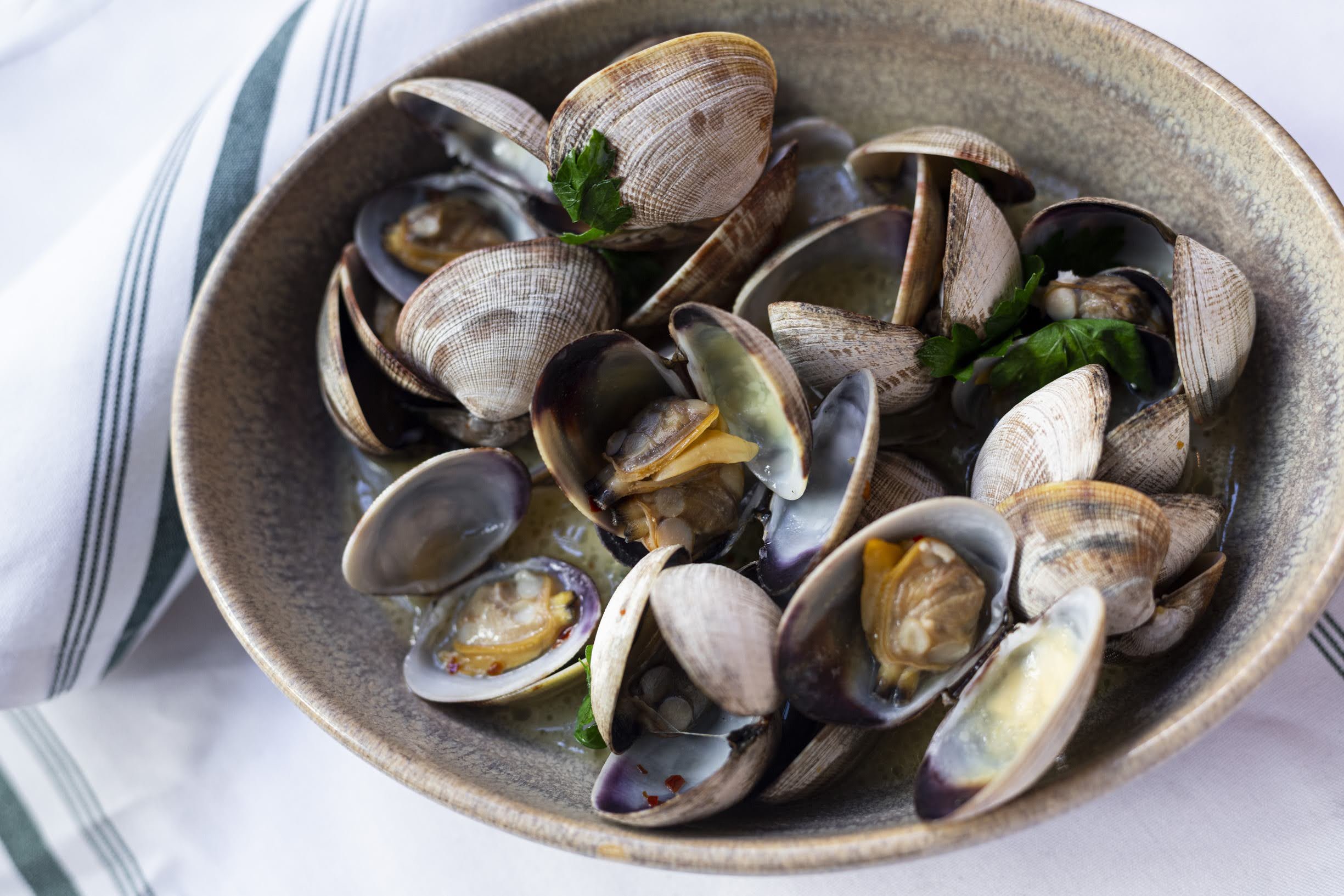 A bowl of cooked steamed clams garnished with fresh parsley on a white and green striped napkin.