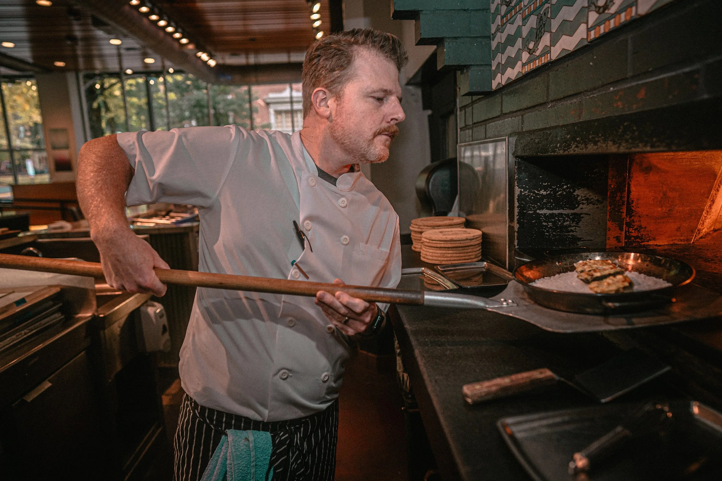 A male chef wearing a white apron and striped pants is using a pizza peel to remove a pizza from a commercial oven. The kitchen has large windows and modern decor.