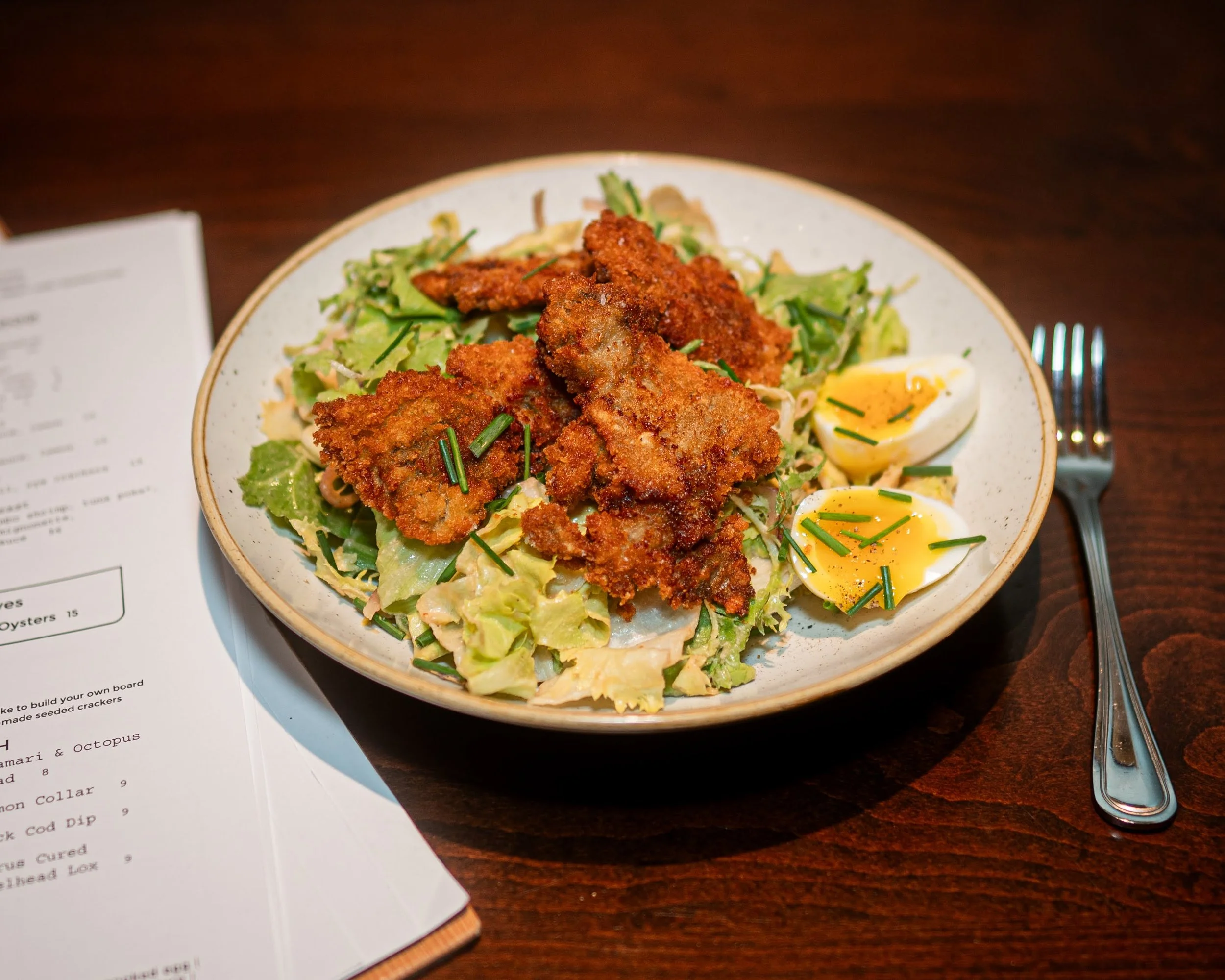 A plate of fried chicken served salad with chopped greens and halved boiled eggs garnished with chives, on a dark wooden table with a fork, knife, and menu nearby.