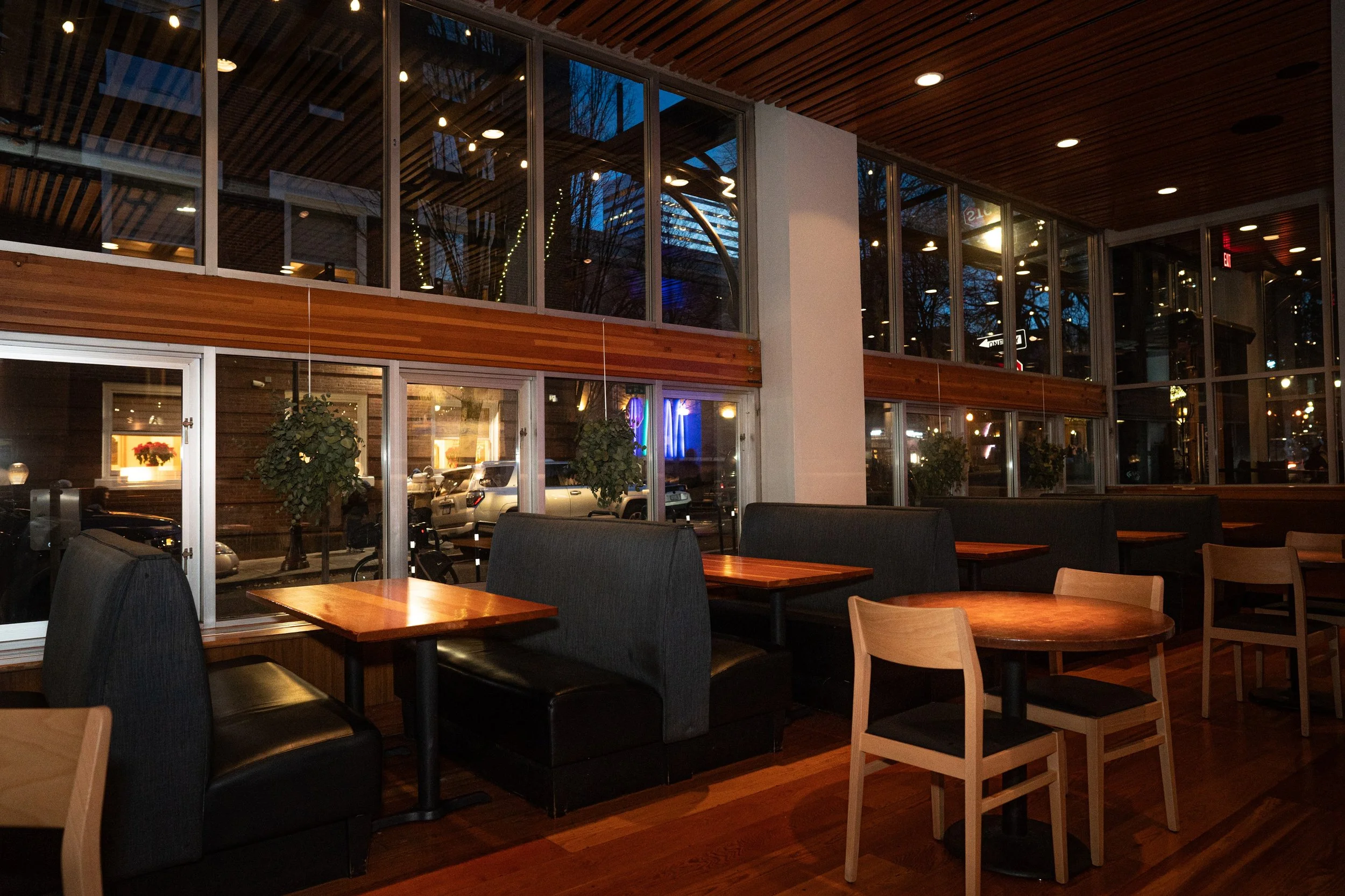 Interior of a modern restaurant with large glass windows, wooden ceiling, and a mix of black and light wood chairs around wooden tables. The evening sky is visible outside through the windows, along with parked cars and trees.
