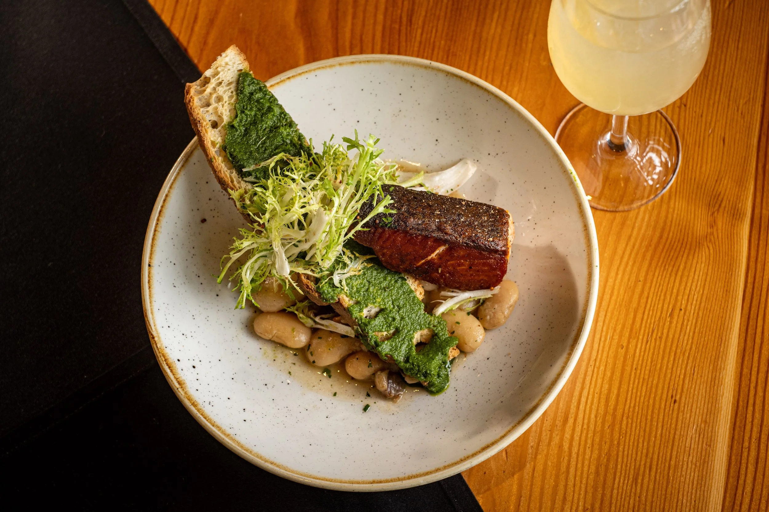 A white ceramic bowl containing white beans, leafy greens, and grilled meat, garnished with microgreens, on a wooden table with a glass of lemonade nearby.