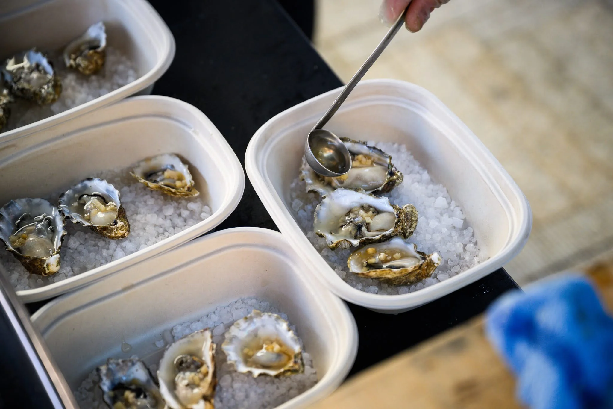 Several oyster shells on a bed of ice in white containers, with a person using a spoon to serve or prepare the oysters.