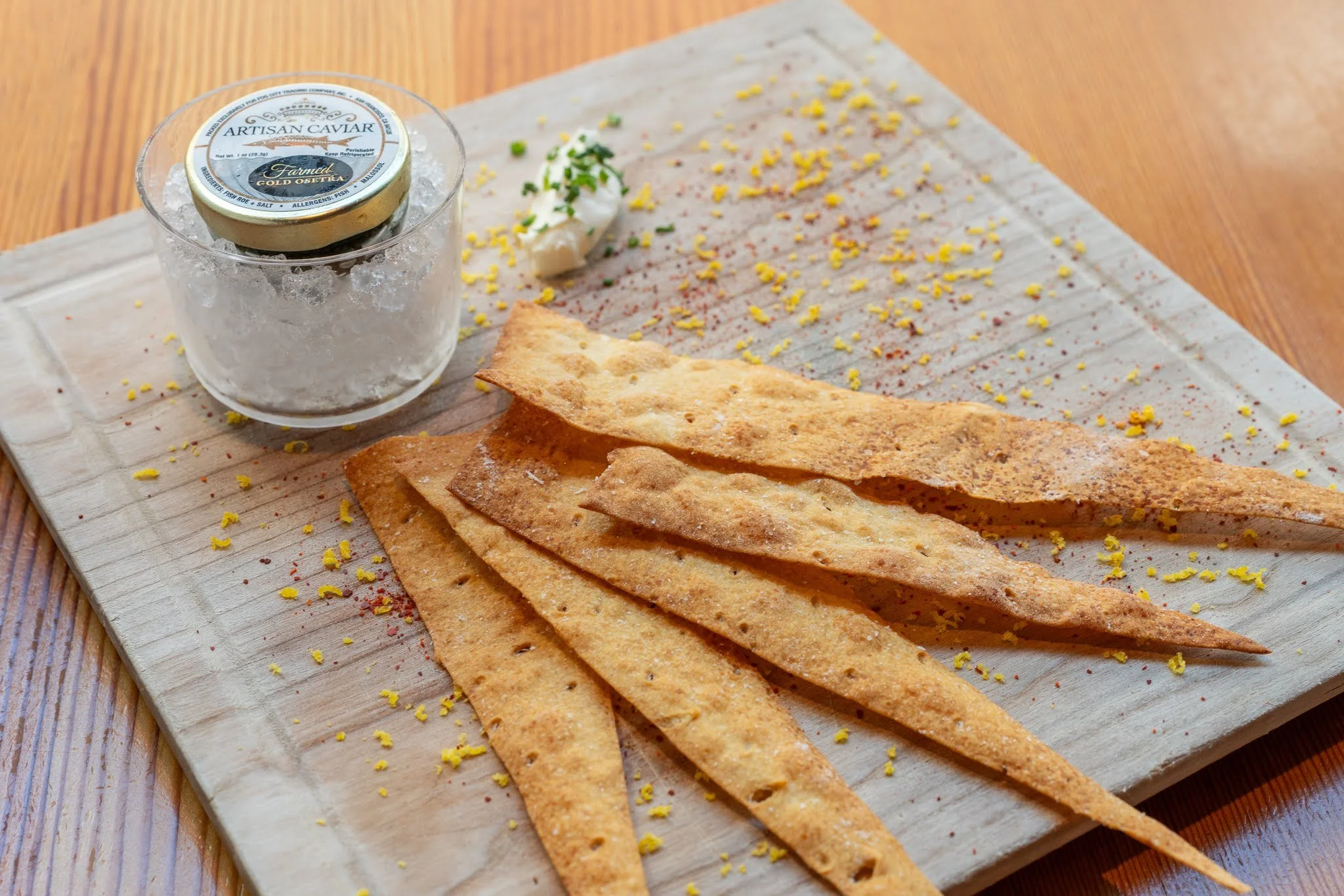 A glass of crushed ice topped with a container of artisan caviar, along with crispy flatbread, garnished with chopped herbs and lemon zest, on a wooden table.