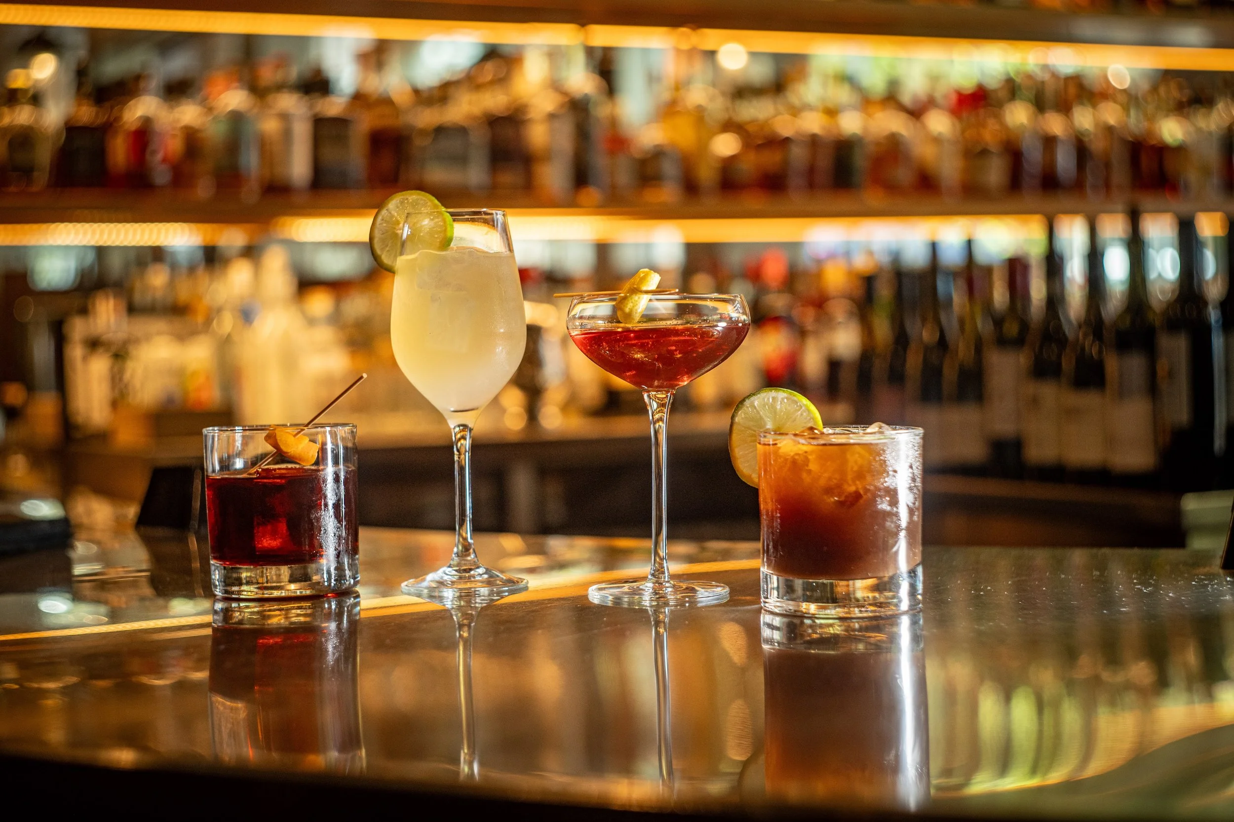 Various colorful cocktails with lime slices on a bar counter, blurred bar shelves in the background.