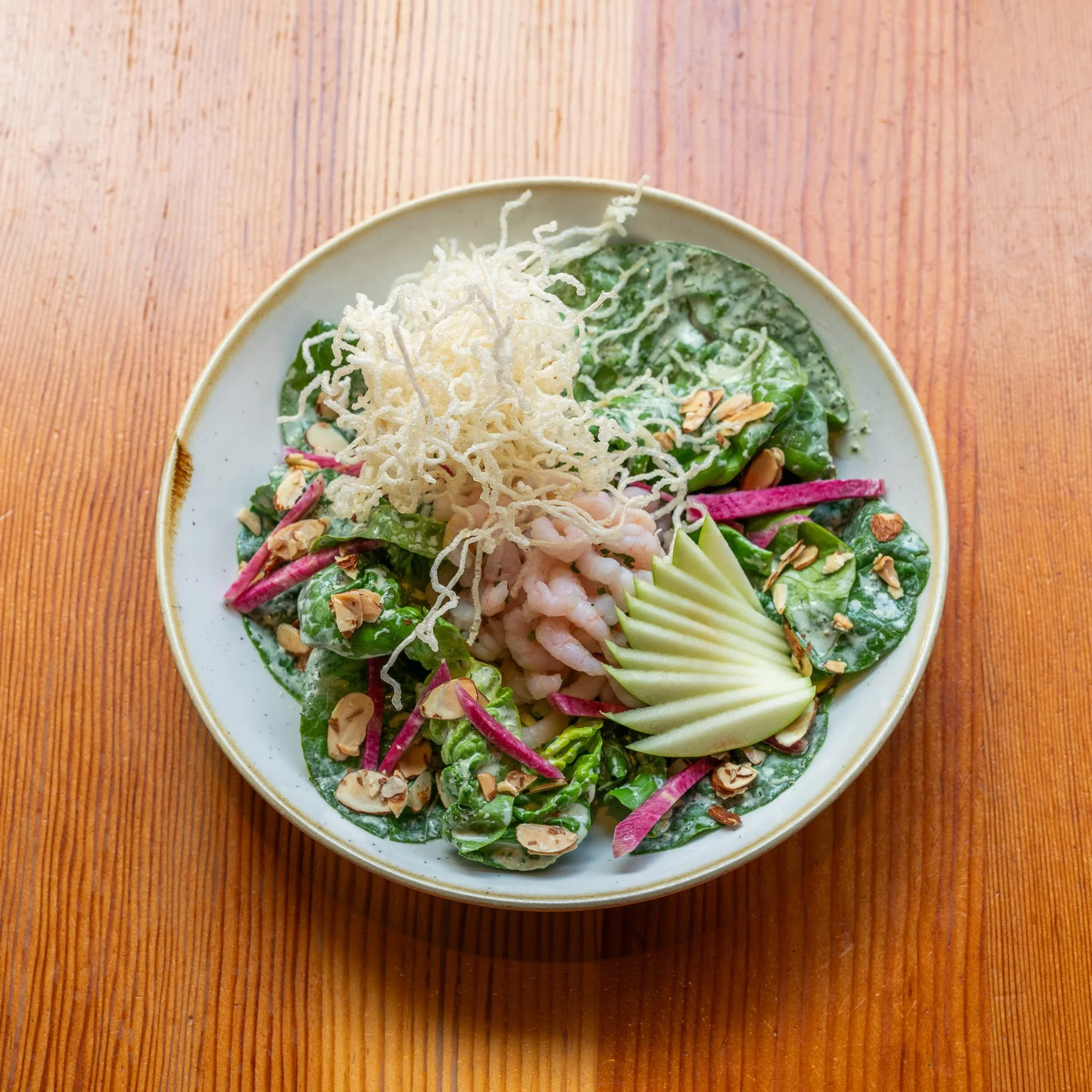A bowl of salad with shredded cheese, sliced almonds, and various greens on a wooden table.