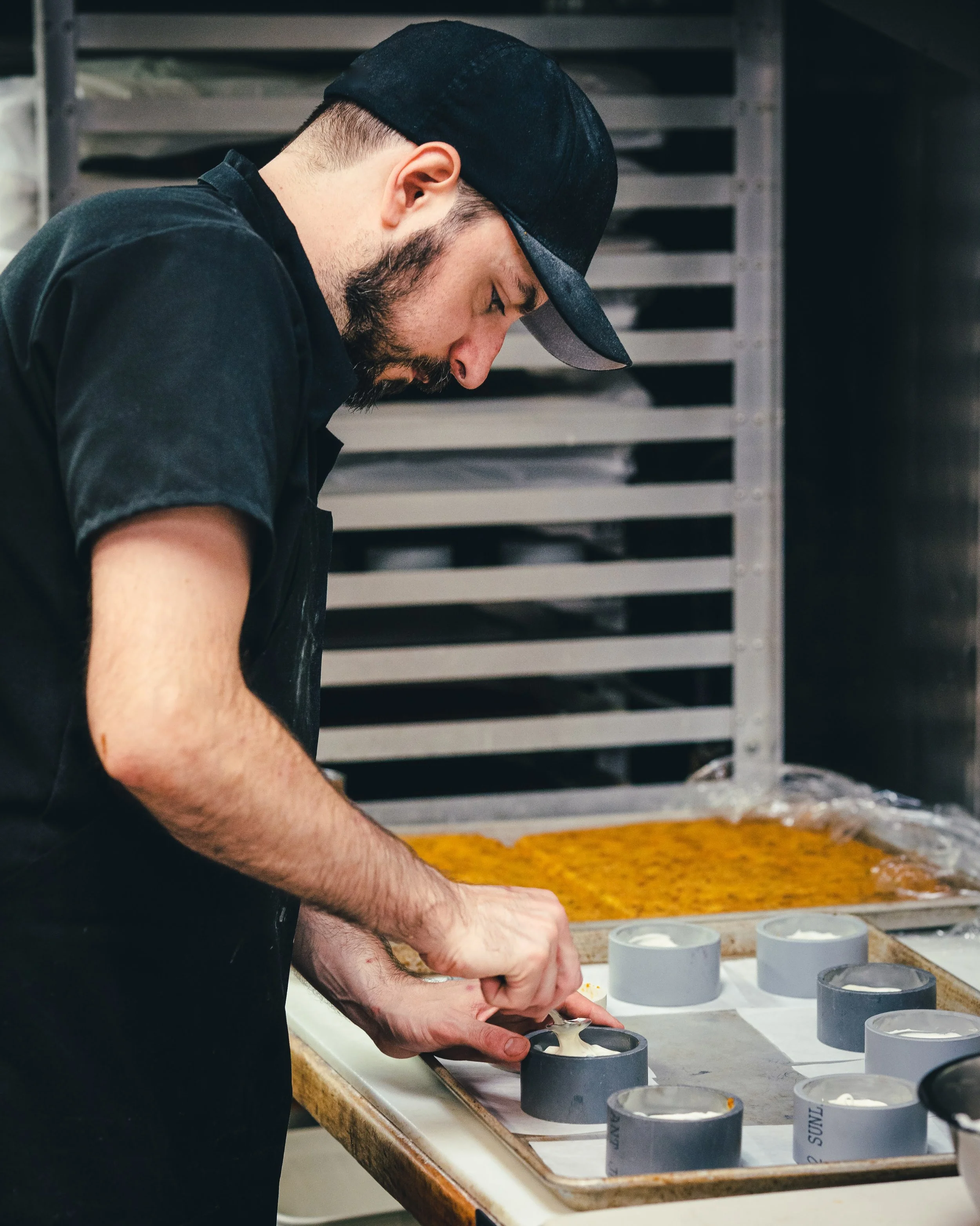 A man with a beard wearing a black cap and black shirt working at a table with cylindrical containers filled with a white substance, possibly ice cream or similar frozen dessert, in a commercial kitchen or production setting.