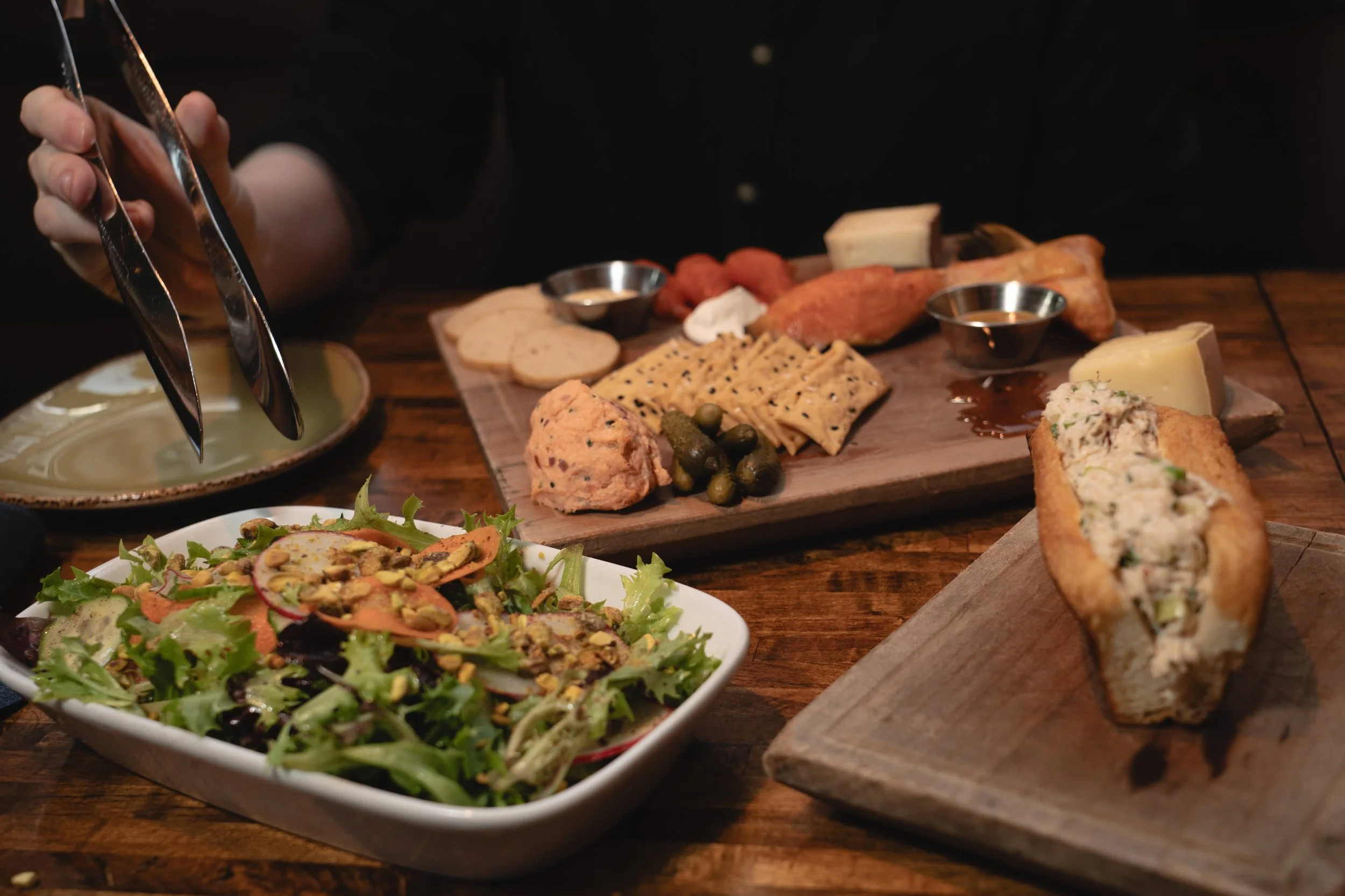 A person uses metal tongs to serve food onto a plate. In the background, a wooden board holds a variety of cheeses, crackers, pickles, and dips. In the foreground, a plate of mixed greens salad with nuts and sliced vegetables is visible.