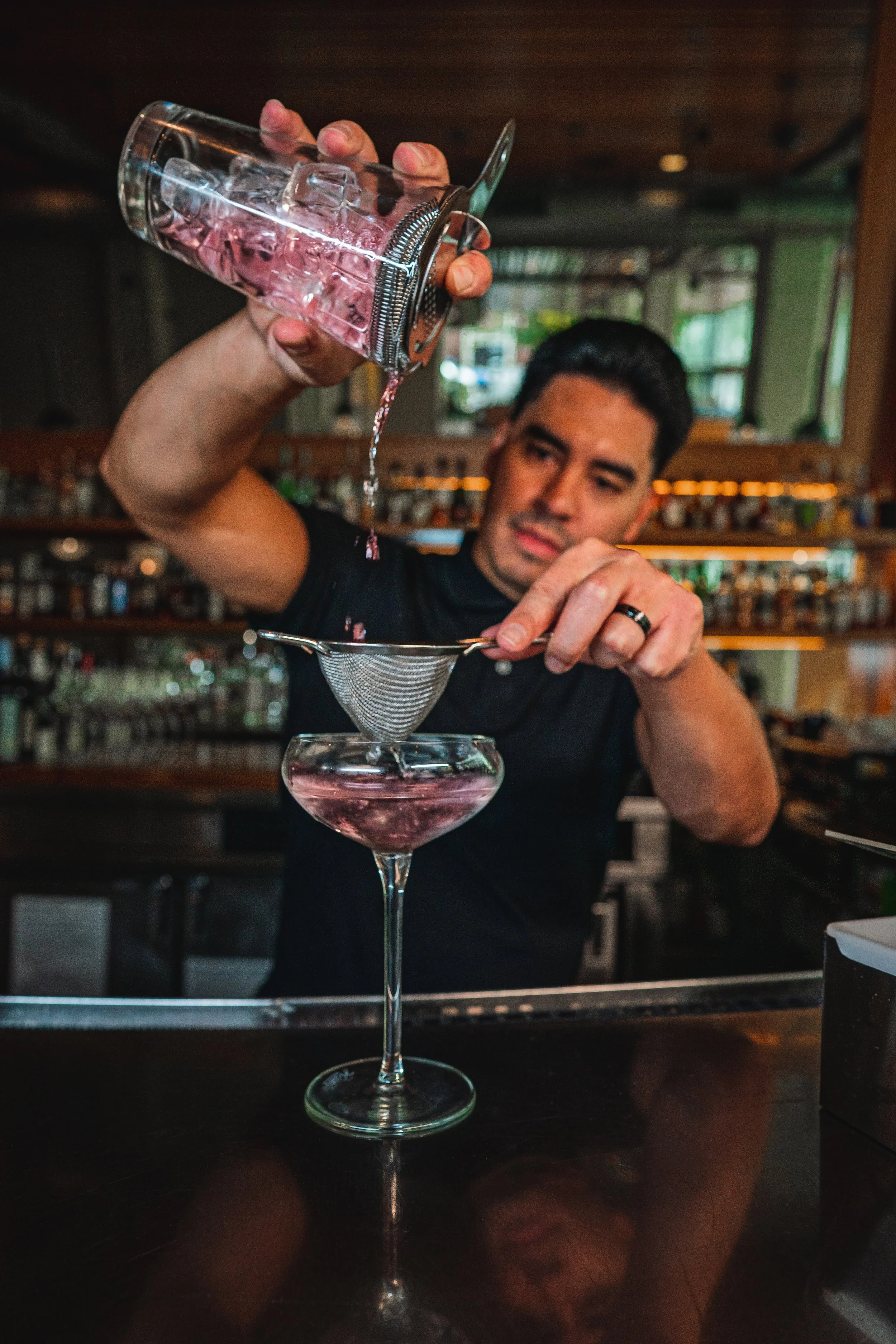 A bartender pours pink liquid through a fine mesh strainer into a cocktail glass, with a bar background.