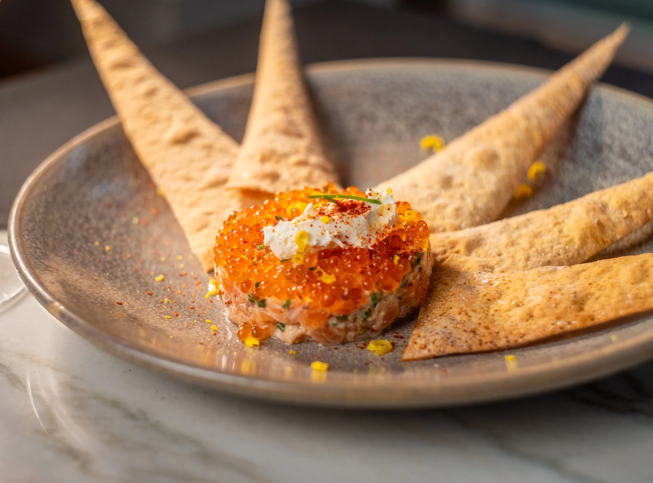 A plate of sushi featuring a piece topped with orange fish roe, garnished with a dollop of white cream, a green herb, and red seasoning, accompanied by crispy rolled chips.