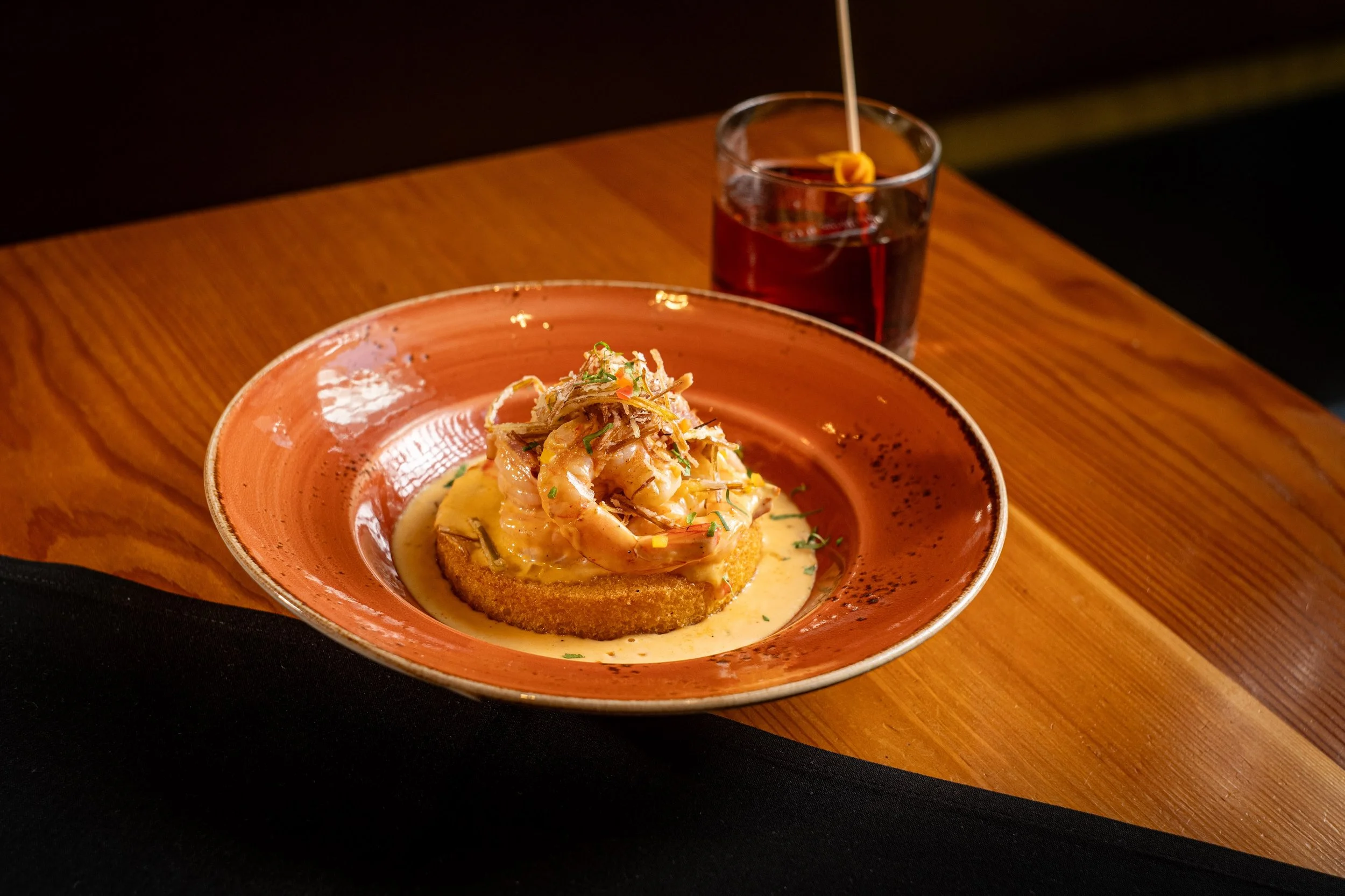A plate of shrimp and vegetables atop a piece of bread with a creamy sauce, accompanied by a glass of red beverage with a lemon slice.