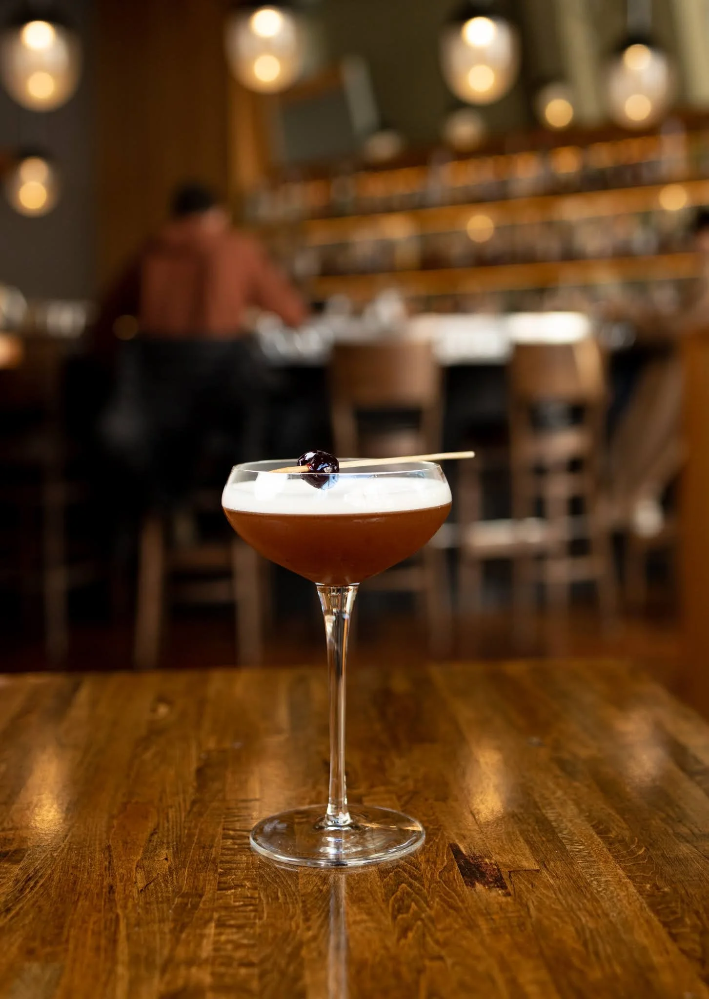 A cocktail in a stemmed glass garnished with a cherry on a wooden table in a dimly lit bar or restaurant with blurred background.
