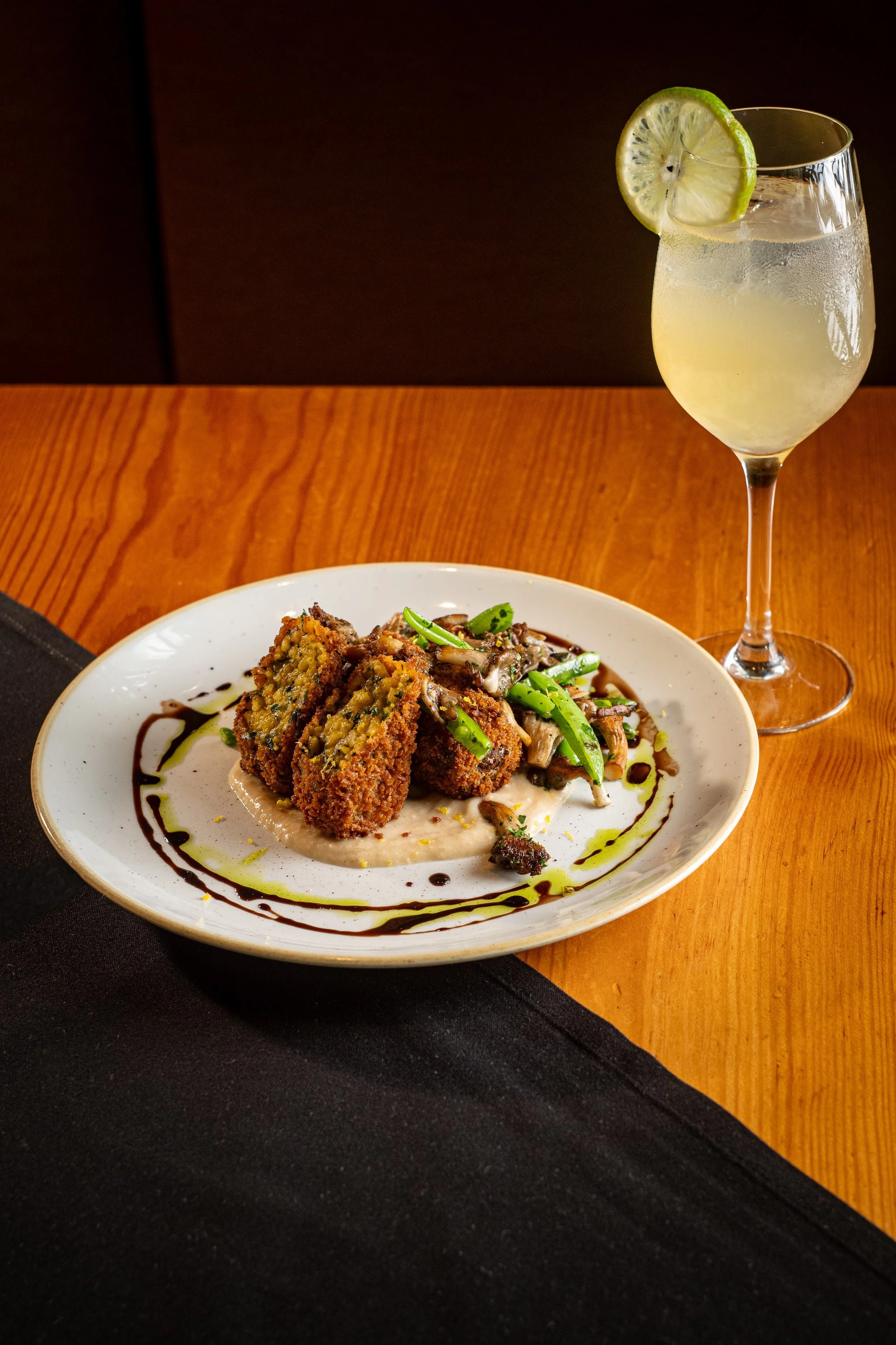 A plate of fried food, possibly fish or chicken, served with a creamy sauce and garnished with green herbs, drizzled with balsamic glaze and olive oil, placed on a wooden table with a black cloth underneath.