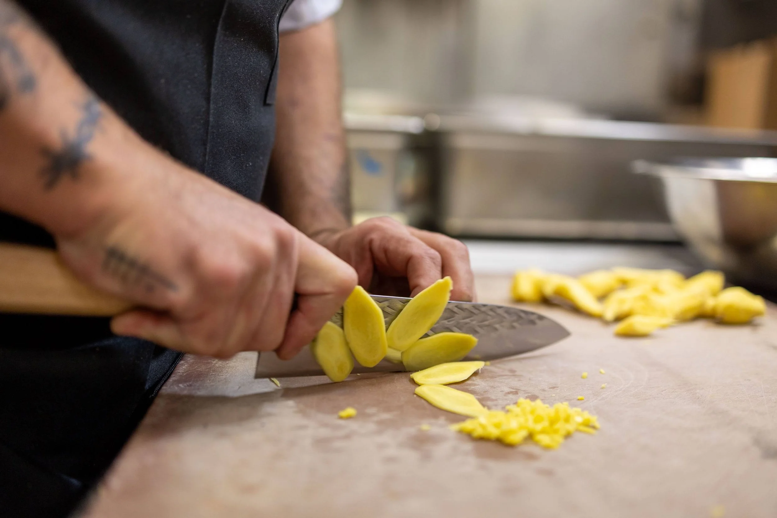 A person slicing yellow vegetables, possibly squash or zucchini, on a wooden cutting board in a kitchen. There are more sliced vegetables on the counter beside the person.