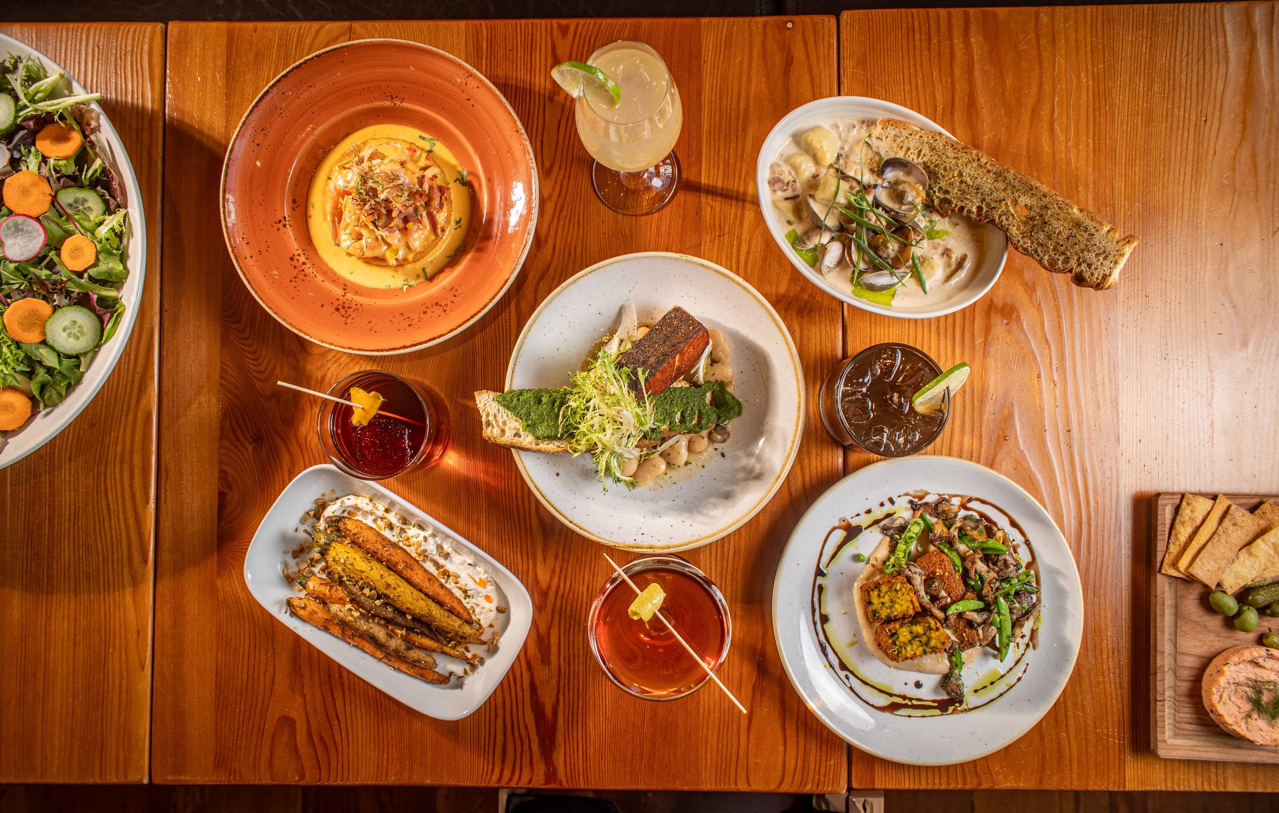 A variety of dishes on a wooden table, including salads, creamy soups, bread, grilled vegetables, and drinks.