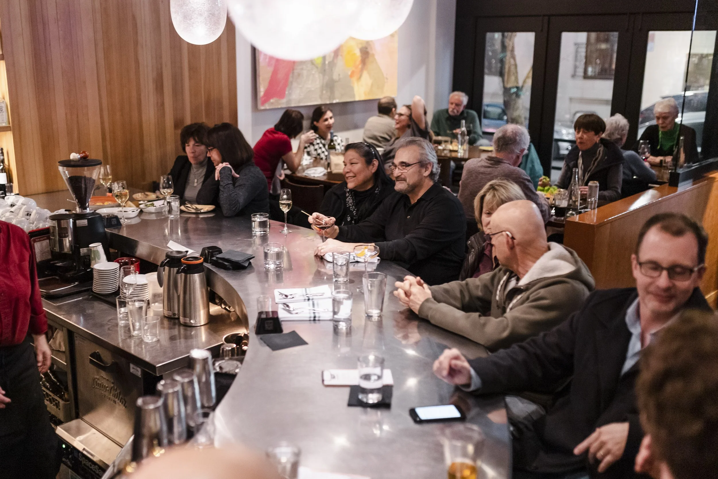 People sitting at a bar counter in a restaurant, chatting and enjoying drinks. There are glasses and bottles on the bar, and some patrons are holding glasses, smiling and engaging in conversation.