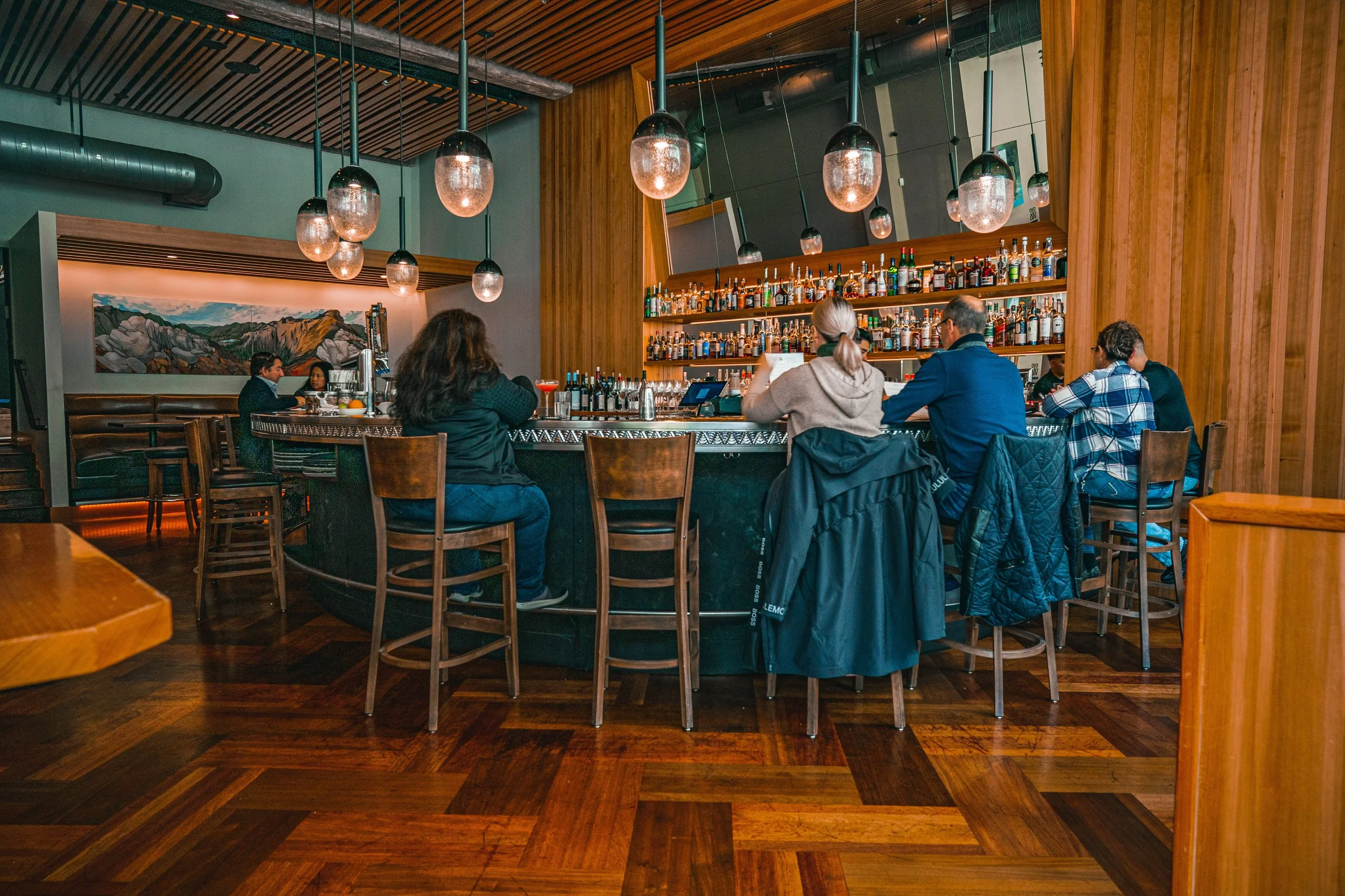 A group of people sitting at a circular bar counter in a modern restaurant or bar with wooden interior and hanging pendant lights.