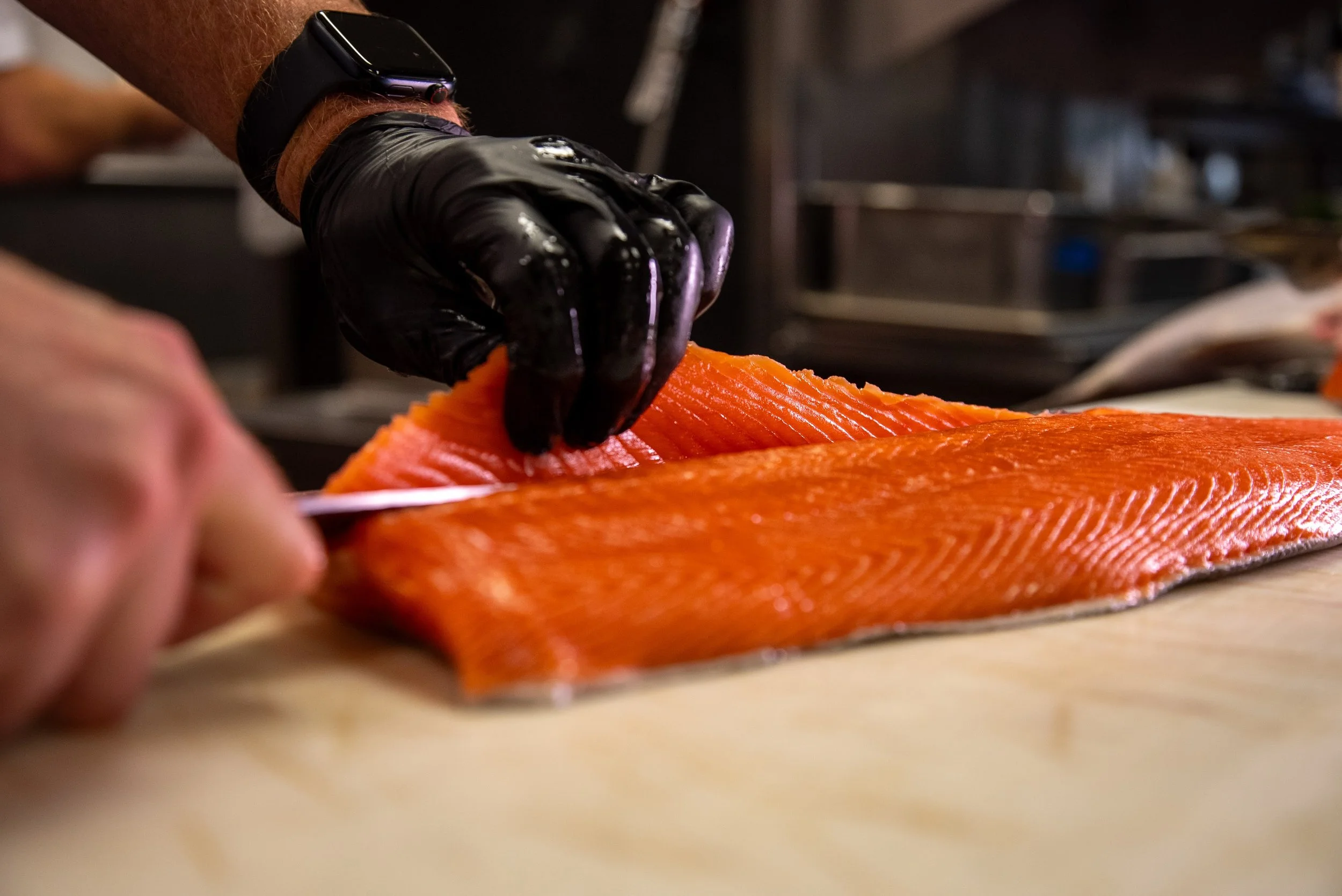 A person wearing a black work glove and a smartwatch slices a piece of orange salmon fish on a cutting board in a kitchen.