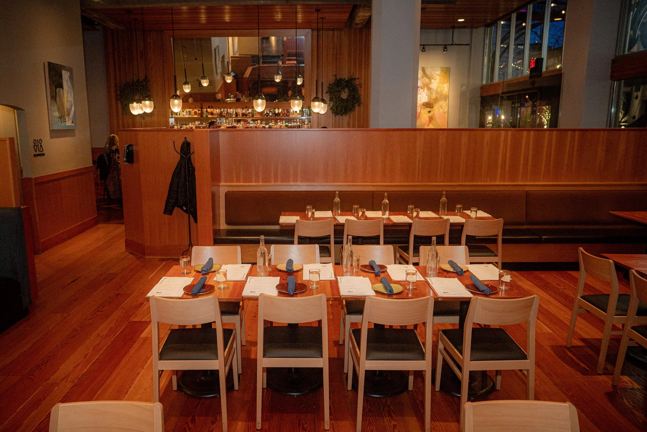 Interior of a restaurant with a long table set for dining, including navy blue napkins, glasses, and plates, with a wooden bar area in the background and modern hanging lights.
