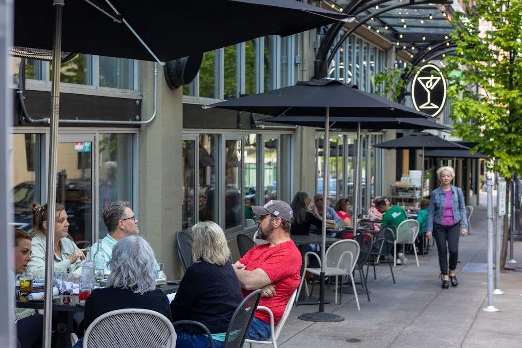 People dining at outdoor café with tables, chairs, and umbrellas on a city sidewalk.