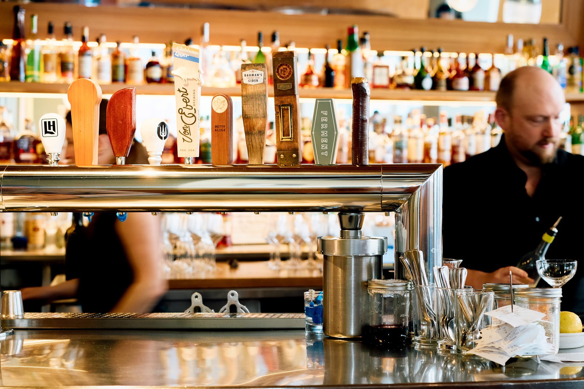 Bar counter with various beer taps, bottles, and glasses, with a bartender smiling in the background.