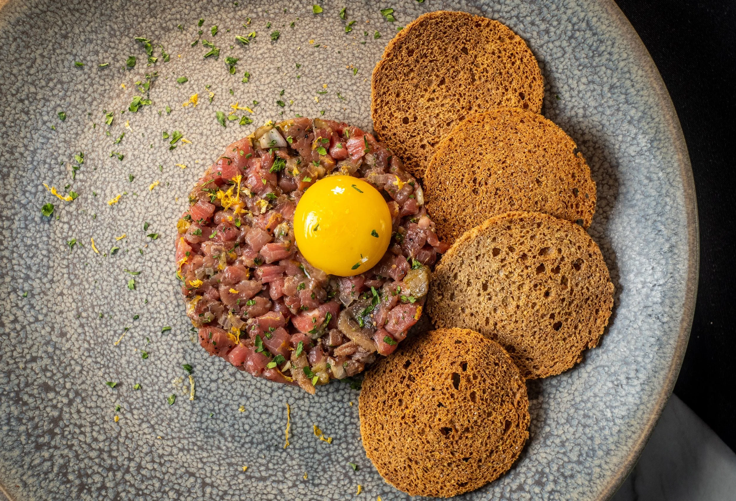 Raw tuna tartare topped with chopped herbs, served with several slices of toasted brown bread on a textured gray plate.