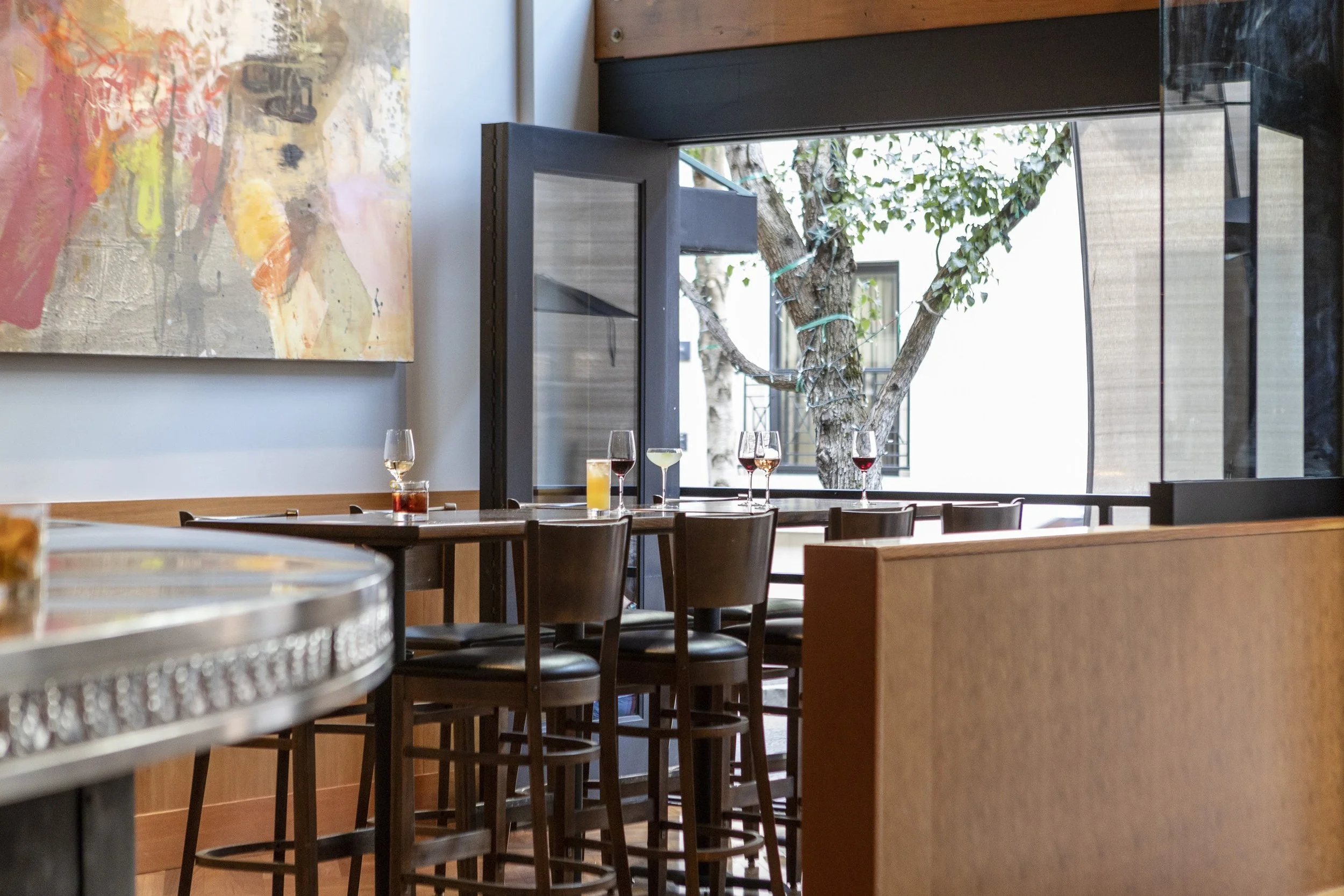 View of a restaurant interior with wooden tables and chairs, colorful abstract art on the wall, and a large open window showing a tree outside with leaves and branches.