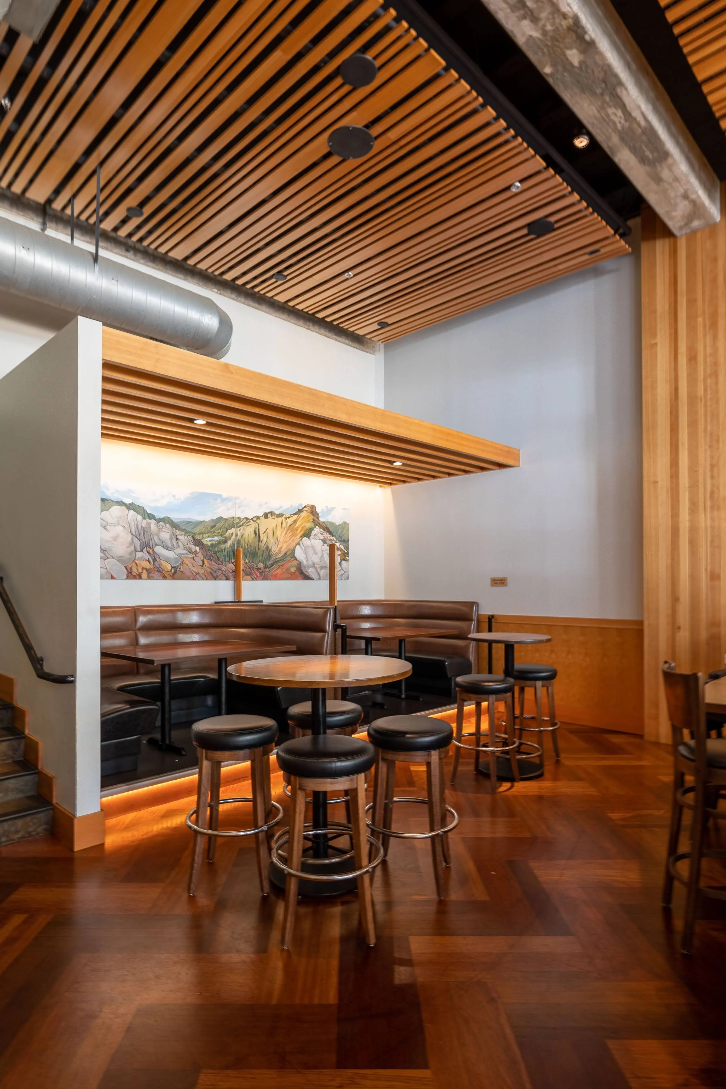 Interior of a modern restaurant or cafe with wooden slatted ceiling, wood paneling, and round tables with stools and cushioned booth seating, and a painting of mountains on the wall.