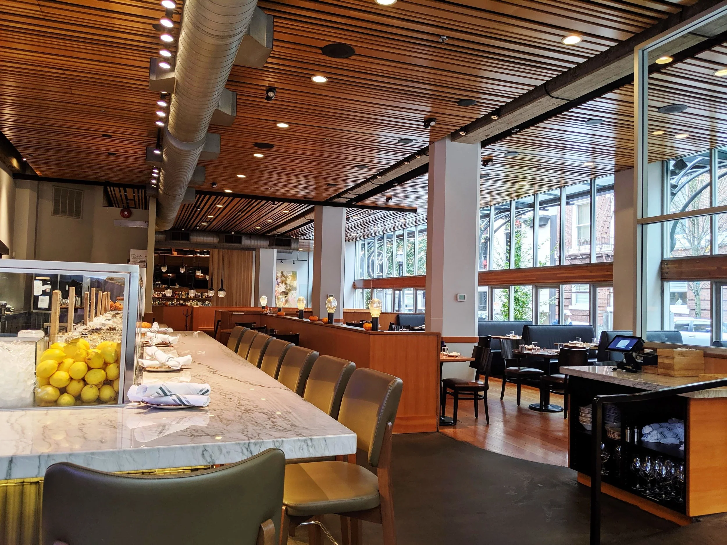 Interior view of a modern restaurant with wooden ceiling, large windows, and a marble countertop with lemons on display.