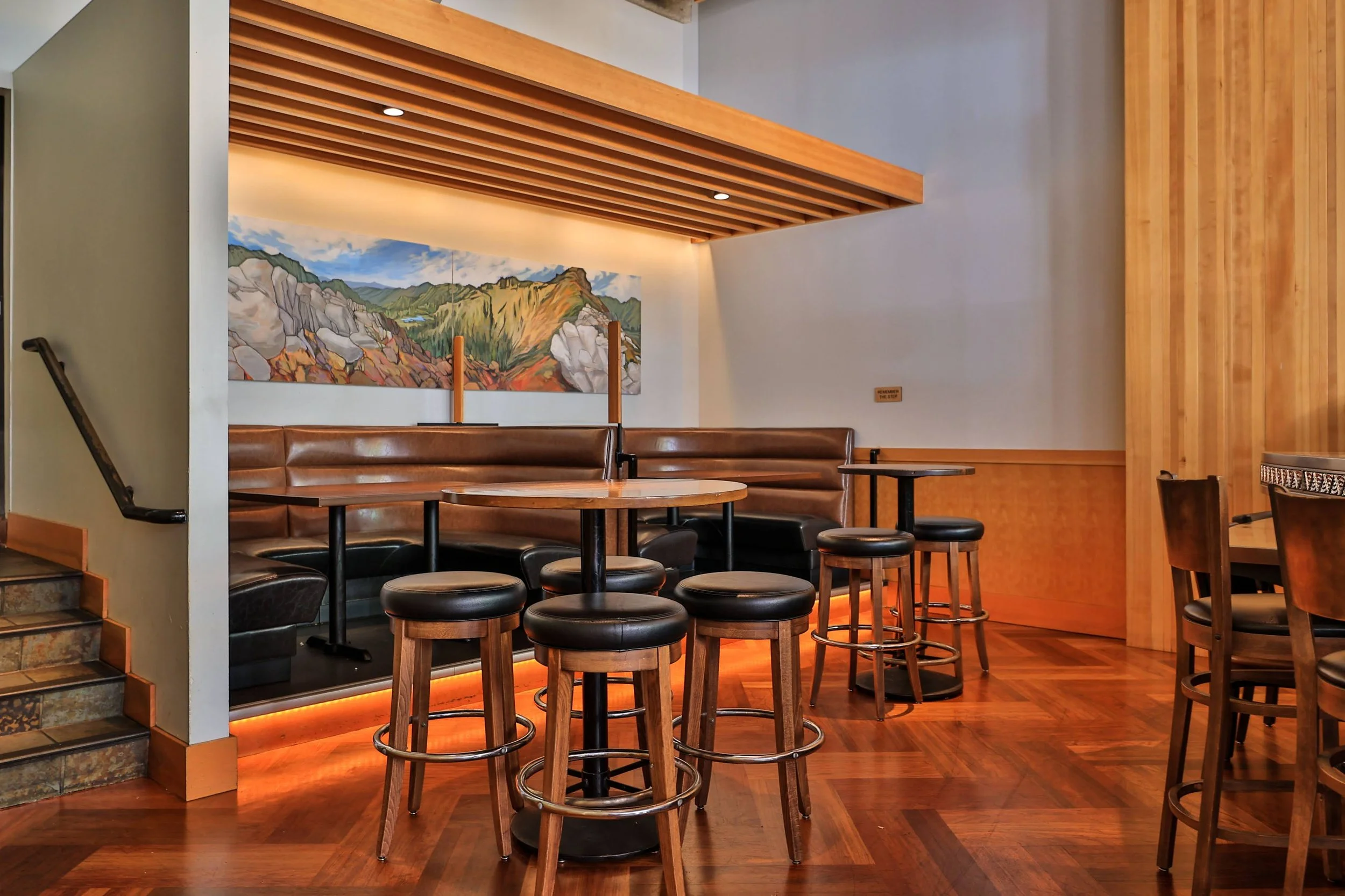 Interior of a cozy restaurant with wooden accents, a mountain mural on the wall, and tables with black chairs and stools.