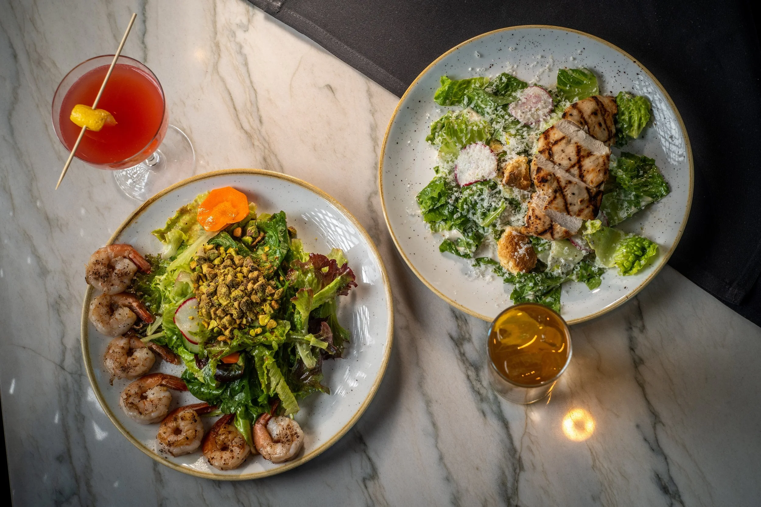Two plates of salad with grilled chicken and shrimp, a glass of pink cocktail with a lemon twist, and a shot of whiskey on a marble table.