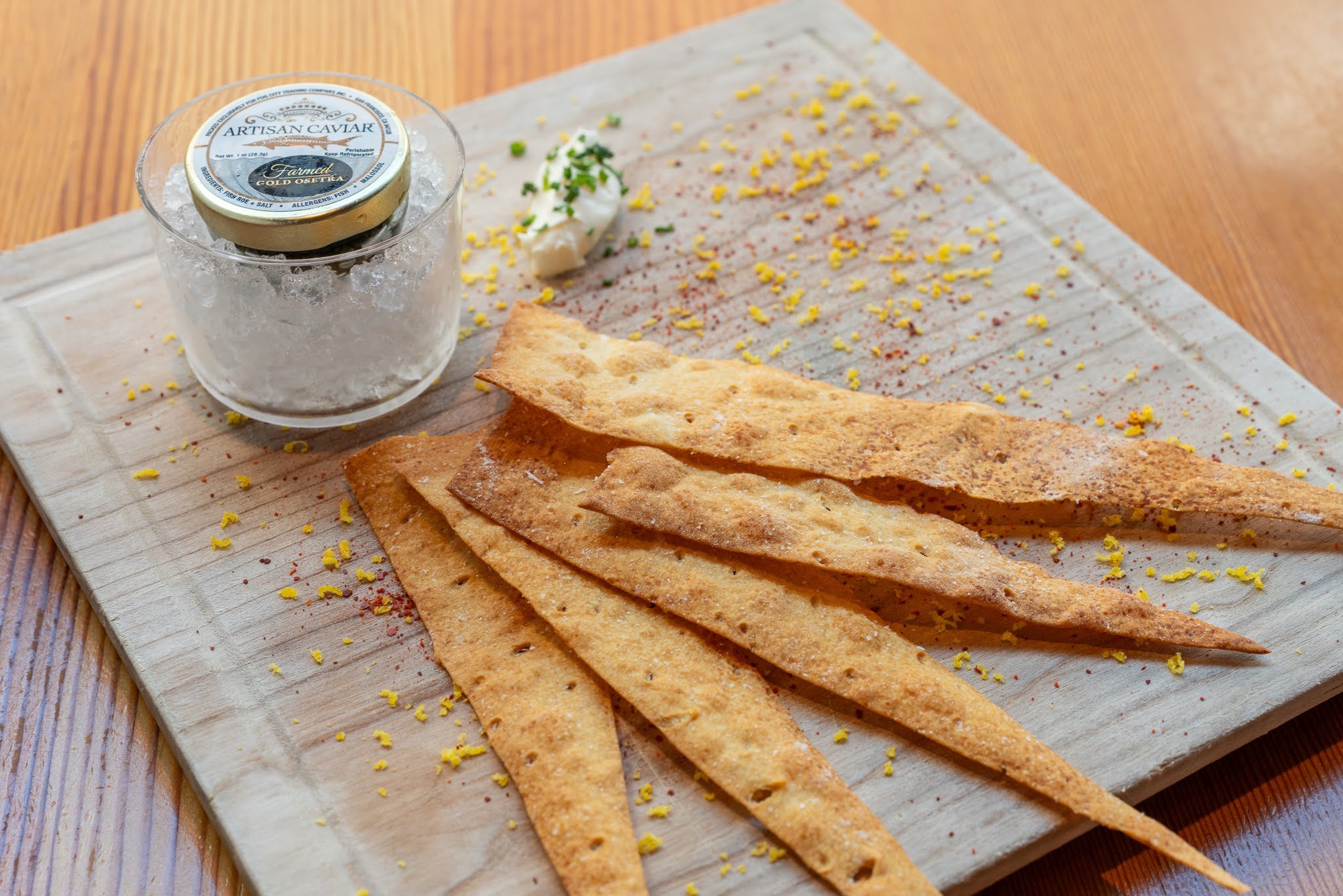 Sliced crispy flatbread with lemon zest, a dollop of sauce garnished with herbs, and a container of artisanal caviar on a wooden cutting board.