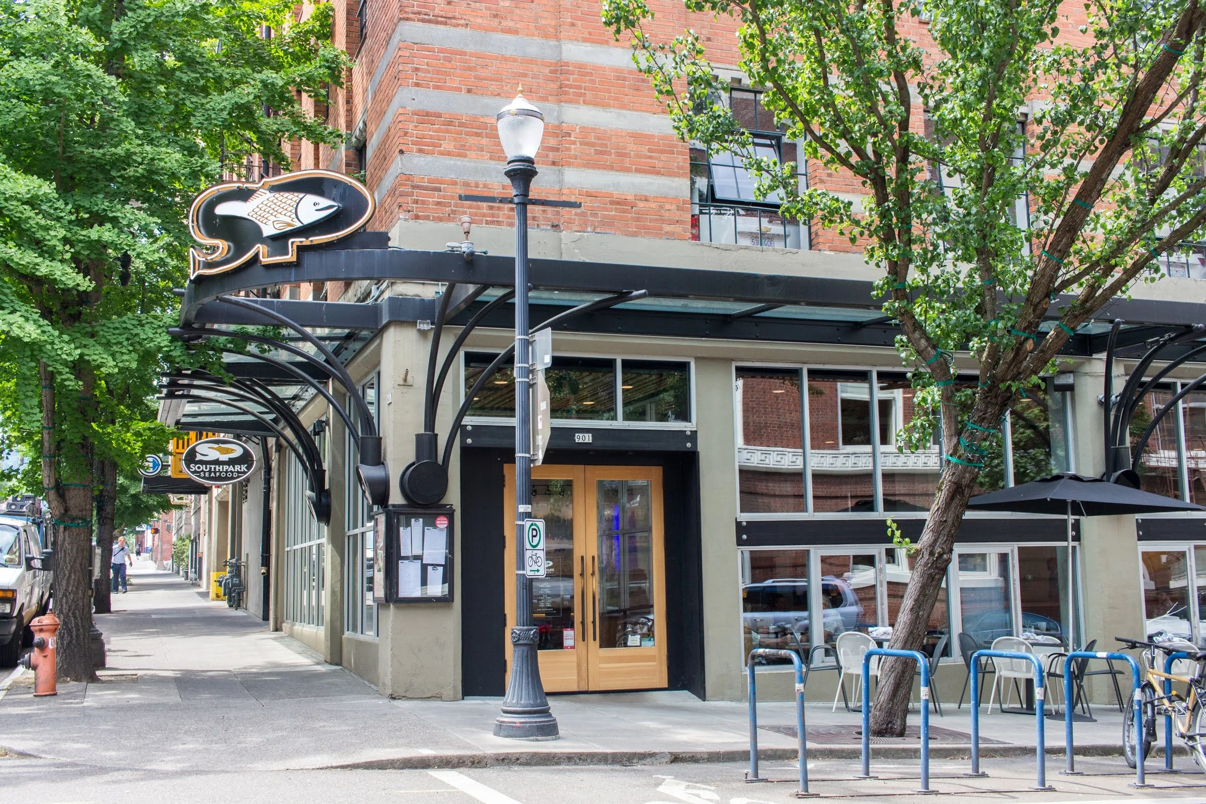 Street view of a restaurant with a fish logo sign, outdoor seating area with metal chairs, bicycles, and trees lining the sidewalk in front. The building has large glass windows and a brick exterior.
