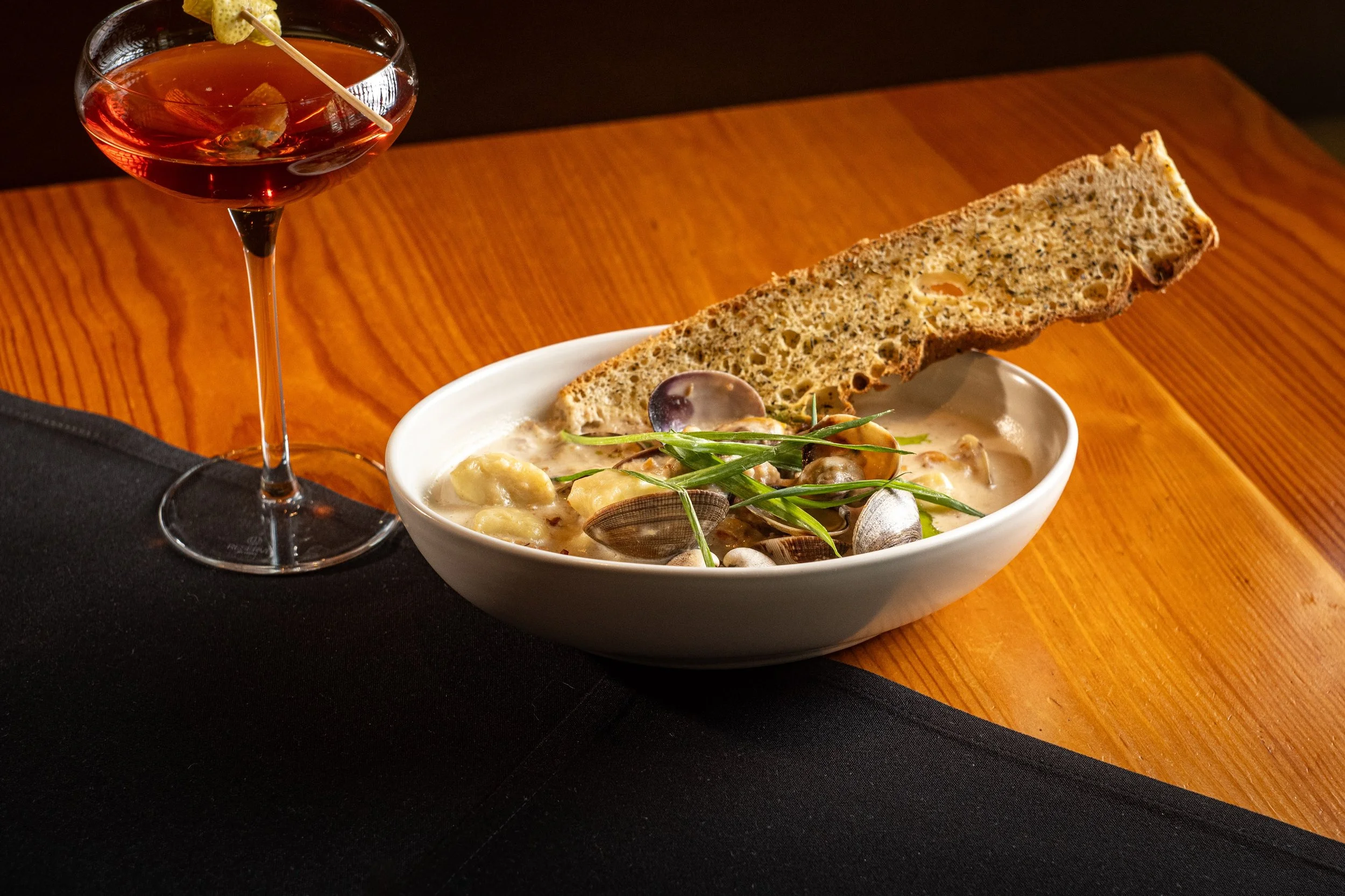 A glass of red cocktail with a lemon twist garnish, and a bowl of creamy seafood chowder with a slice of whole grain bread on a wooden table.